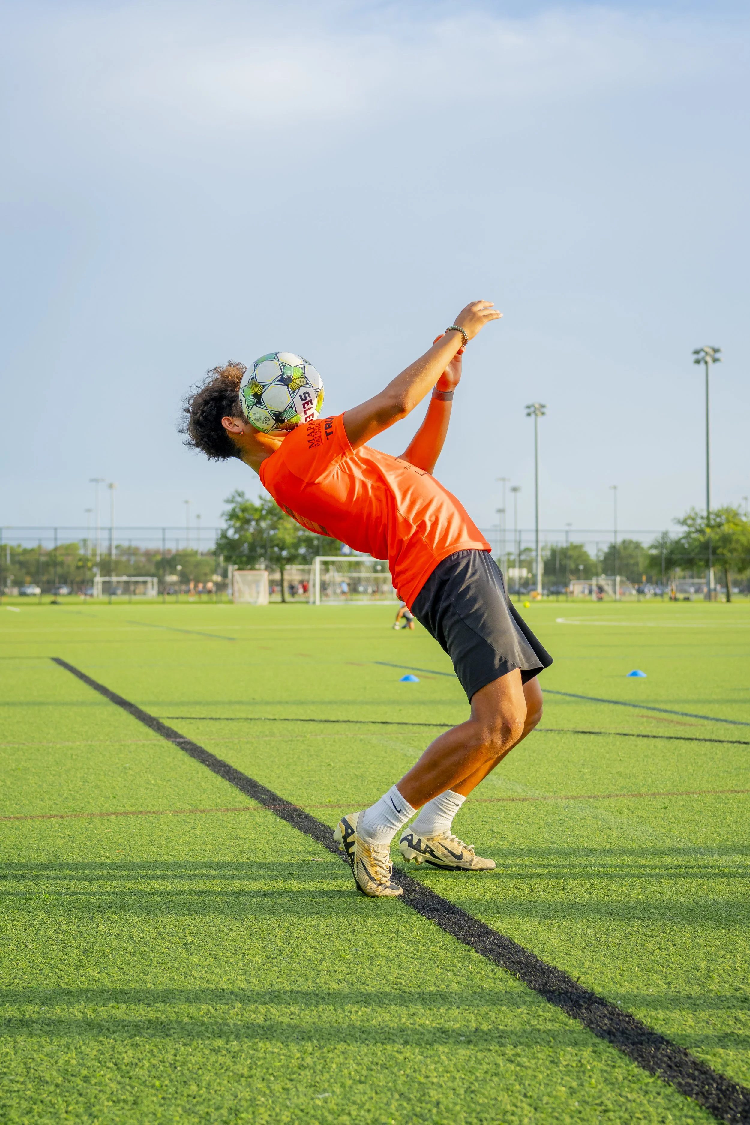 A soccer player in an orange shirt and black shorts is mid-air, heading a soccer ball during practice on a green field with goal posts in the background.