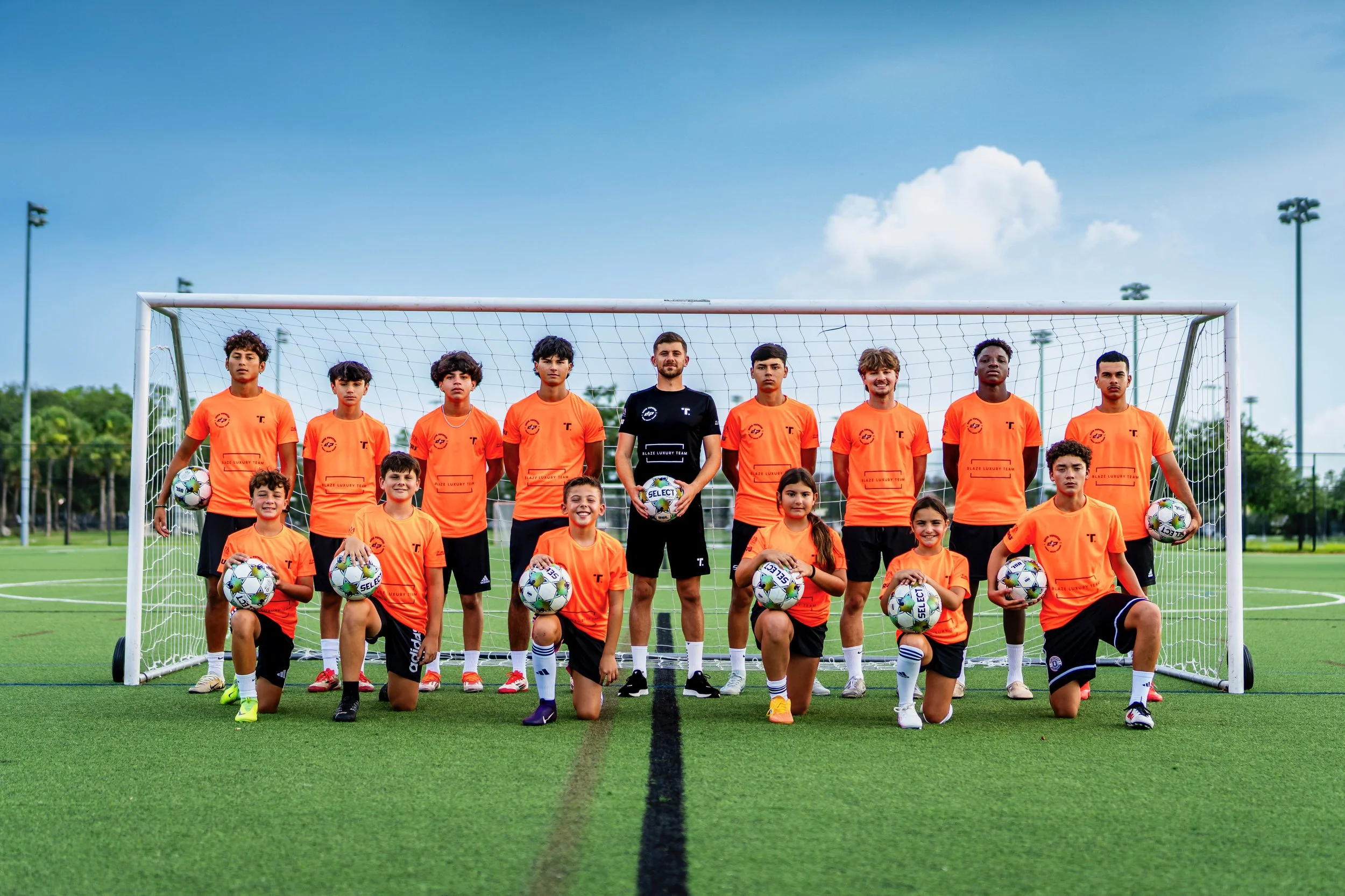 A youth soccer team featuring boys and girls in orange jerseys and black shorts, posing on a soccer field with a coach in the center holding a soccer ball, under a blue sky with clouds.