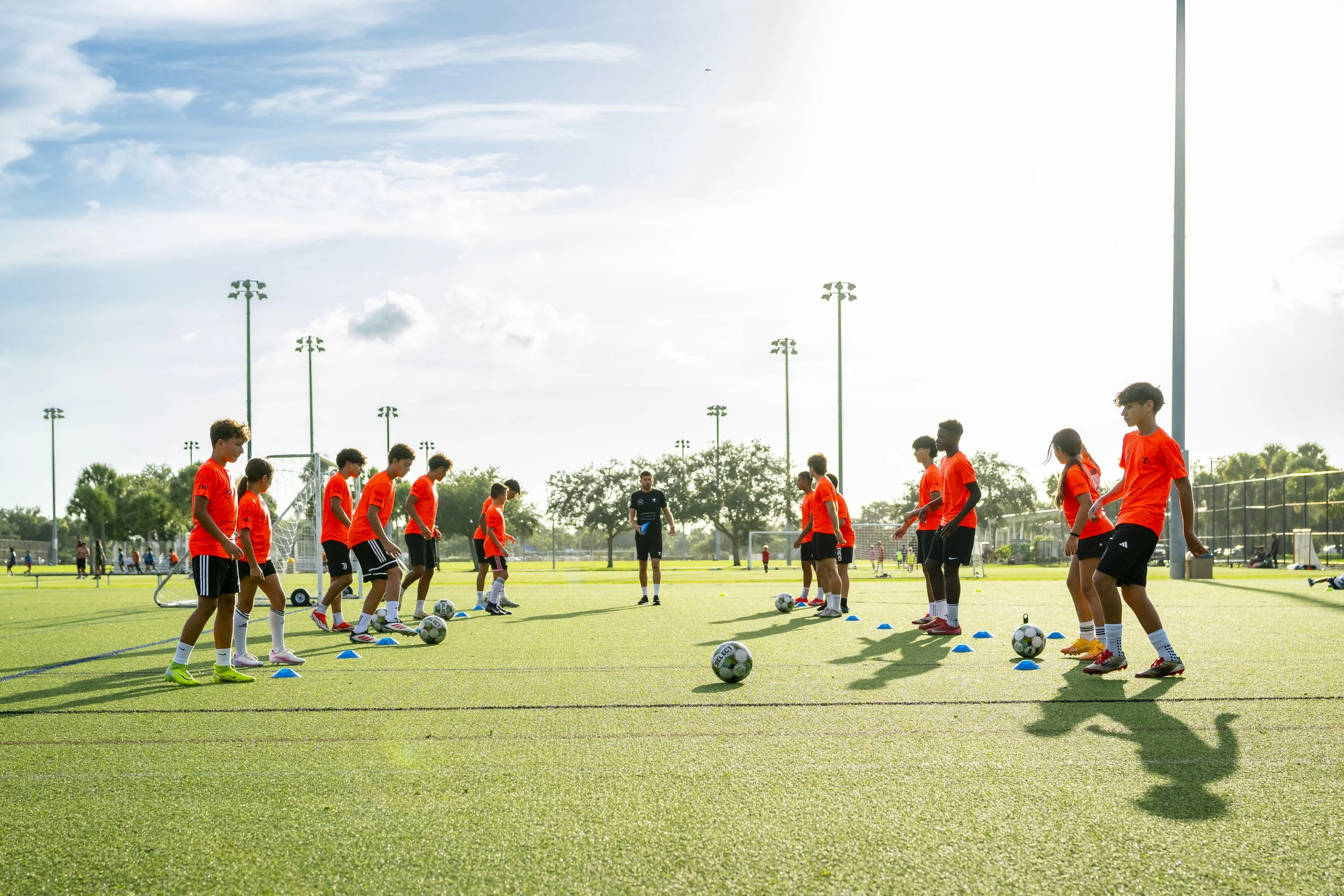 Young soccer players in orange jerseys practicing on a green field with coach, blue cones, and soccer balls under a bright sky.