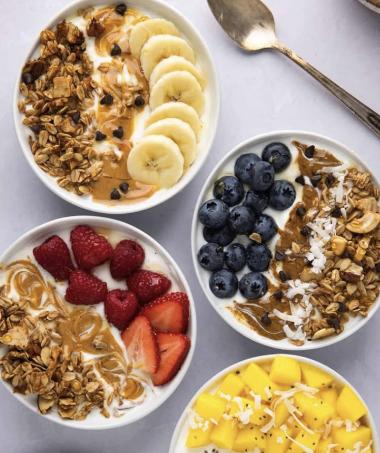 Four bowls of yogurt with various toppings including sliced bananas, blueberries, strawberries, mango, granola, coconut flakes, and drizzle of caramel.