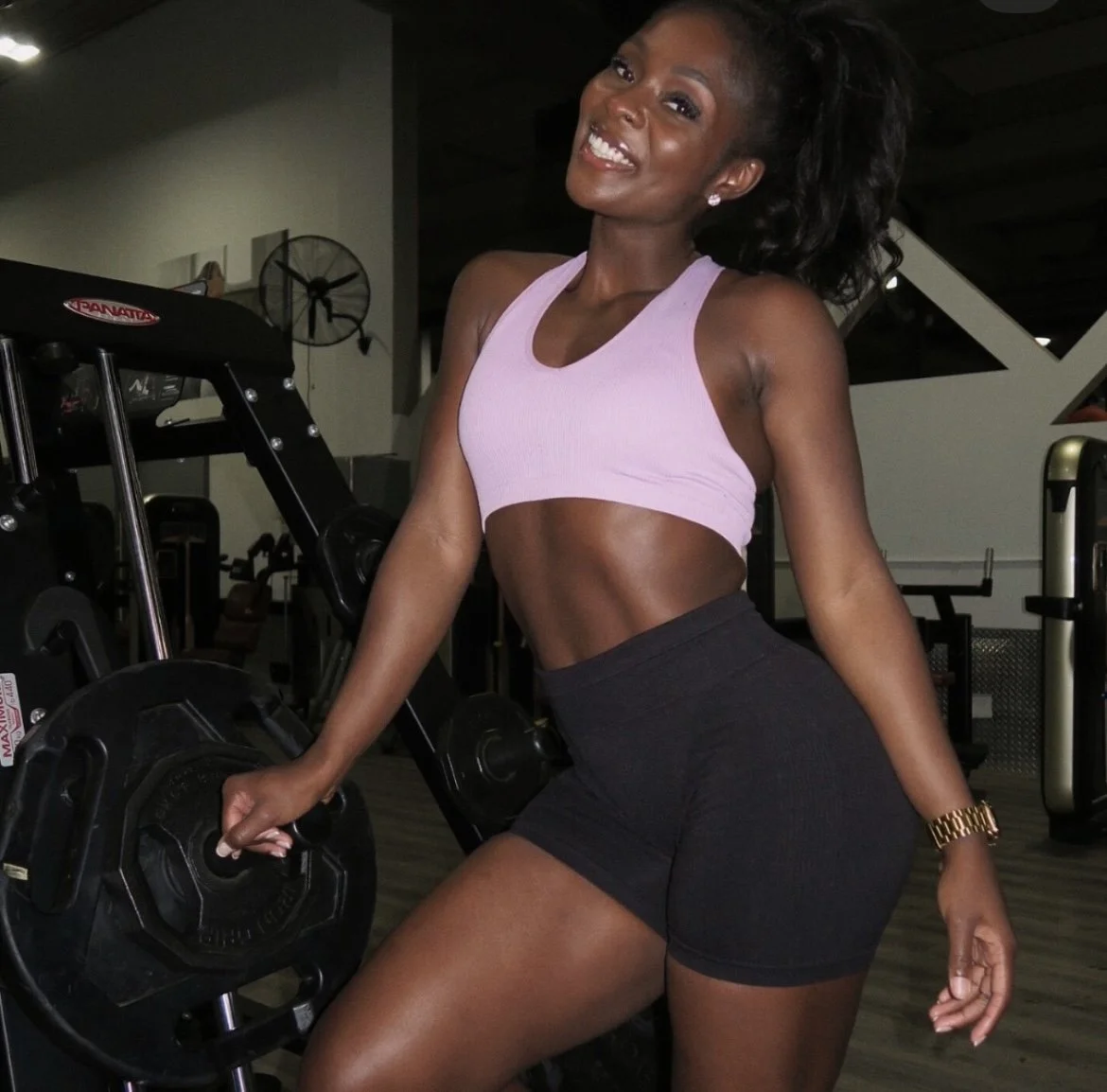 Young woman in workout attire smiling at gym with weightlifting equipment in background.