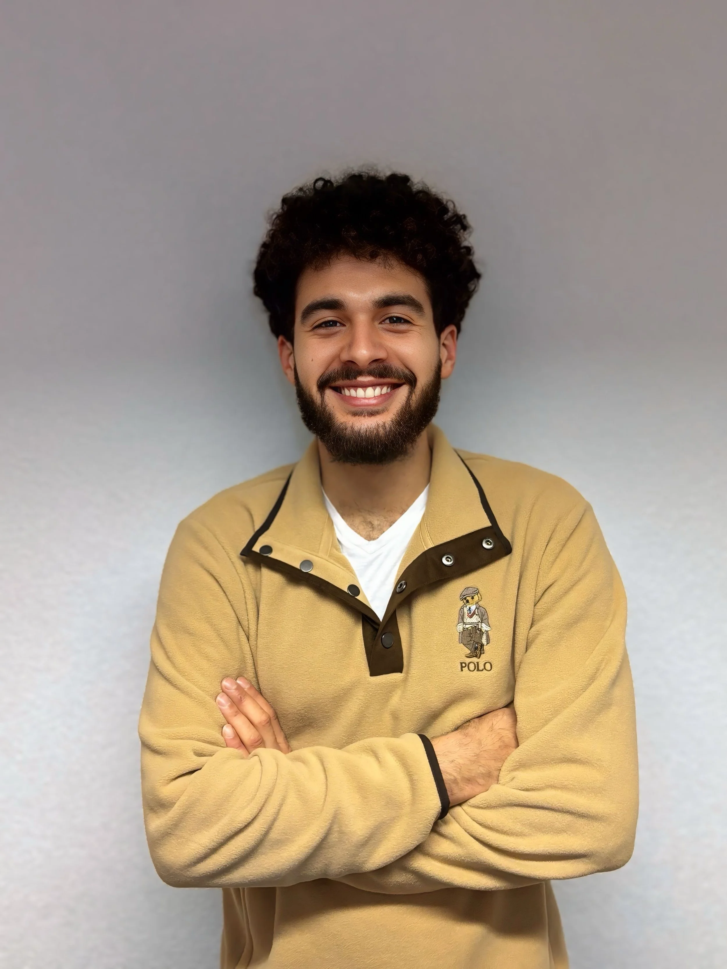 Ein lachender Mann mit dunklen, lockigen Haaren und Vollbart steht vor einer neutralen Wand, trägt ein beige Polo-Hemd mit einem kleinen Polo-Logo auf der Brust, seine Arme sind vor der Brust verschränkt.