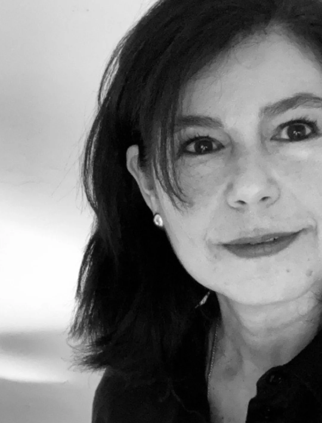 Retrato en blanco y negro de una mujer con cabello oscuro, ojos claros, cejas definidas, usando aretes y sonrisa sutil.