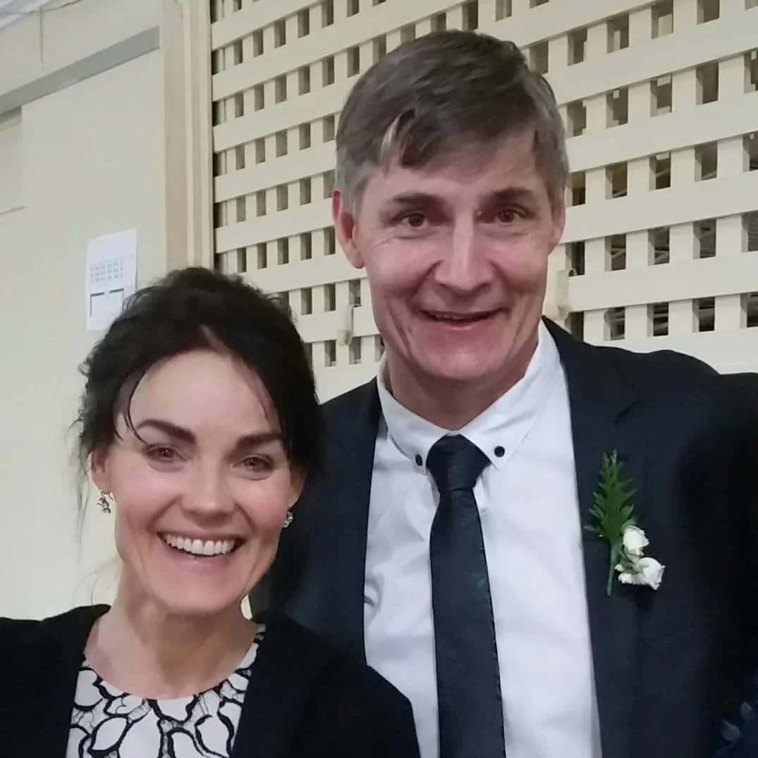 A smiling woman with earrings standing next to a smiling man wearing a suit and tie, with a boutonniere, at a social event with a wooden lattice backdrop.