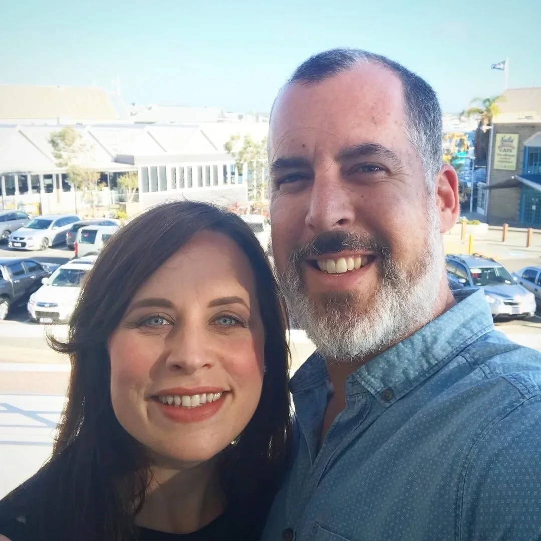 A smiling couple taking a selfie outdoors with a parking lot and buildings in the background.
