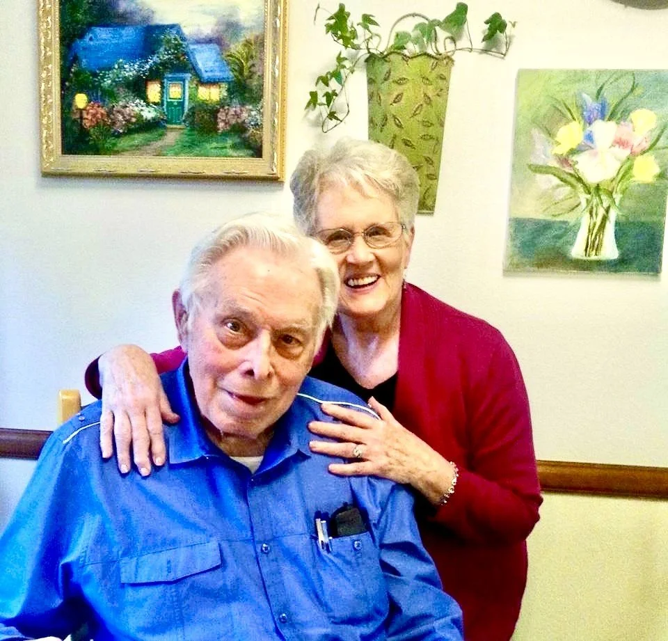 An elderly man and woman smiling, with the woman hugging the man from behind. They are indoors, with paintings and a decorative plant in the background.