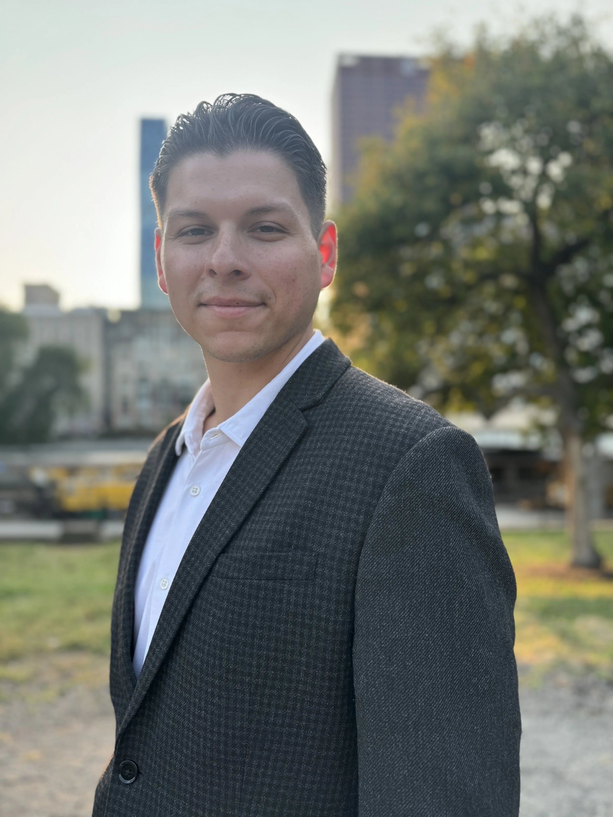 A young man in a dark blazer and white shirt standing outdoors in a city park, with buildings, trees, and a sunset in the background.