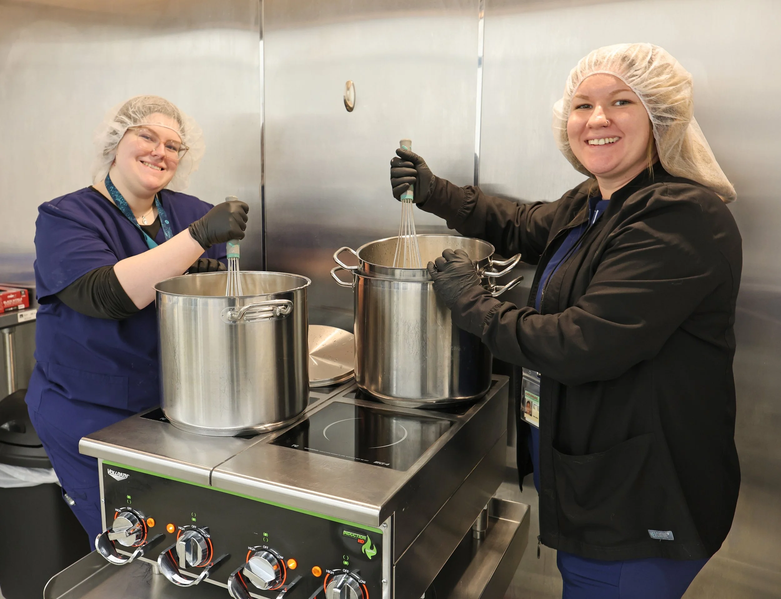 Two women in chef hats and black gloves stirring large stainless steel pots in a commercial kitchen.