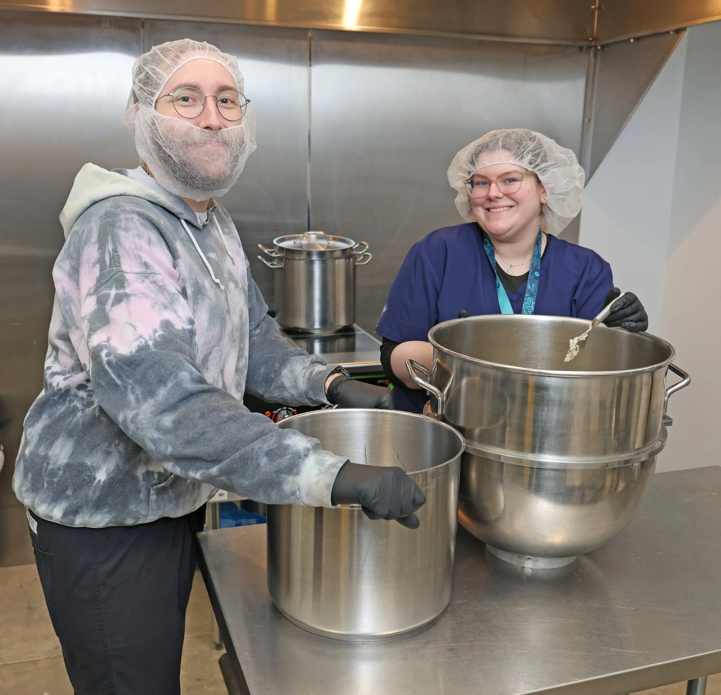 Two people are preparing food in a commercial kitchen. They are wearing hair nets, gloves, and casual clothing. One person is opening a large metal pot while the other is stirring or mixing in the same pot. Both are smiling at the camera.