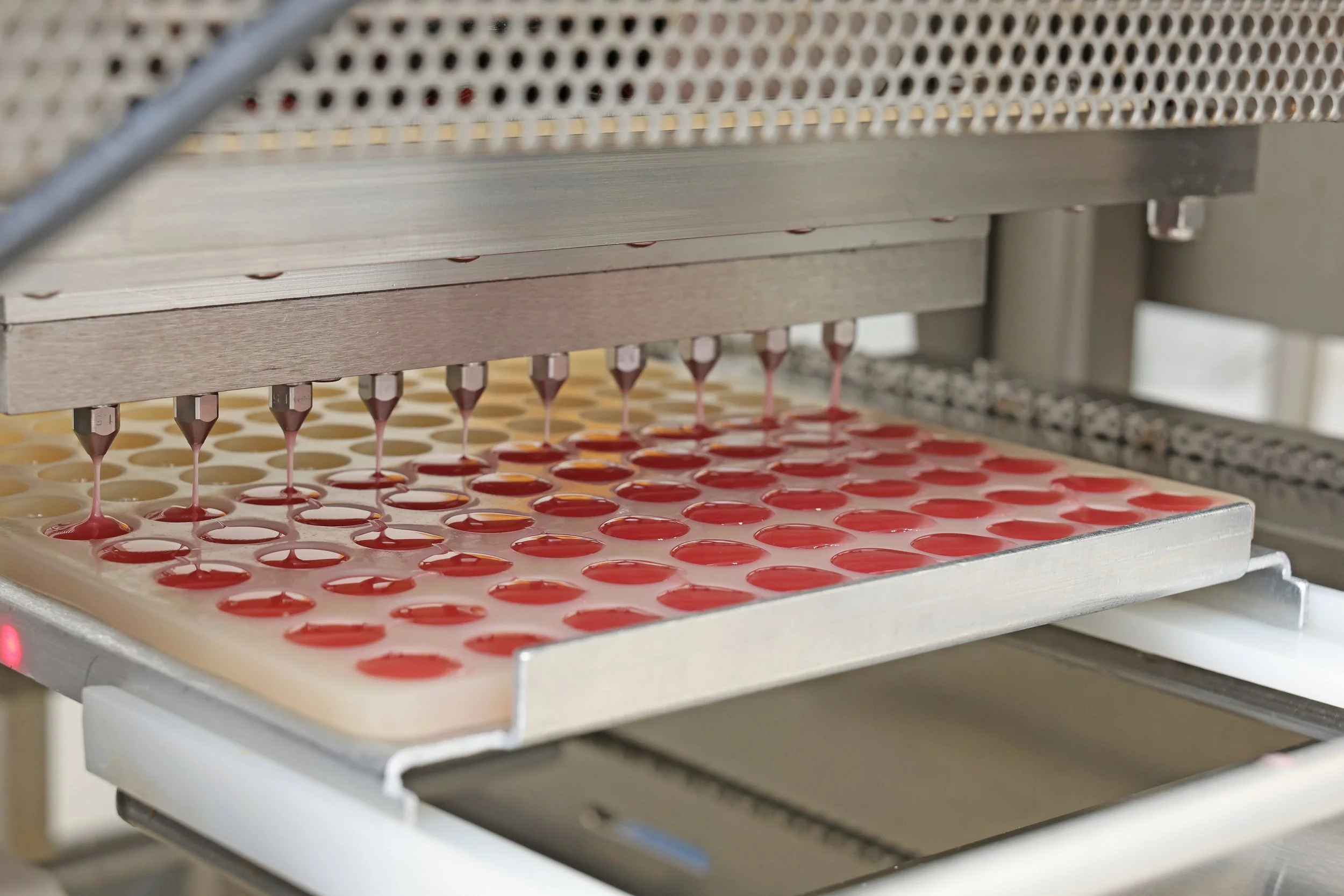 Automated machine dispensing red and yellow goo into circular molds on a conveyor belt in a manufacturing facility.