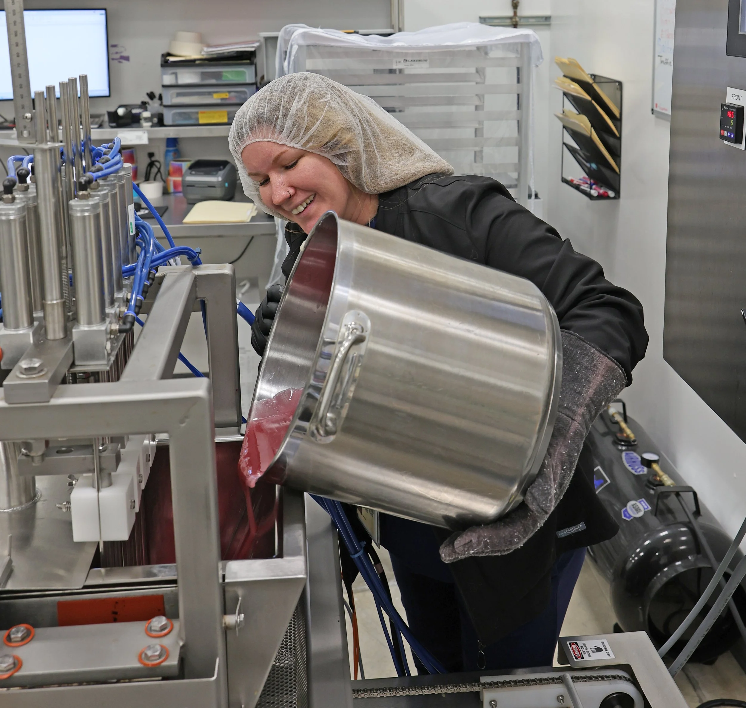 A woman in a lab coat, hair net, and gloves pours a red liquid into a machine in a laboratory or manufacturing setting.