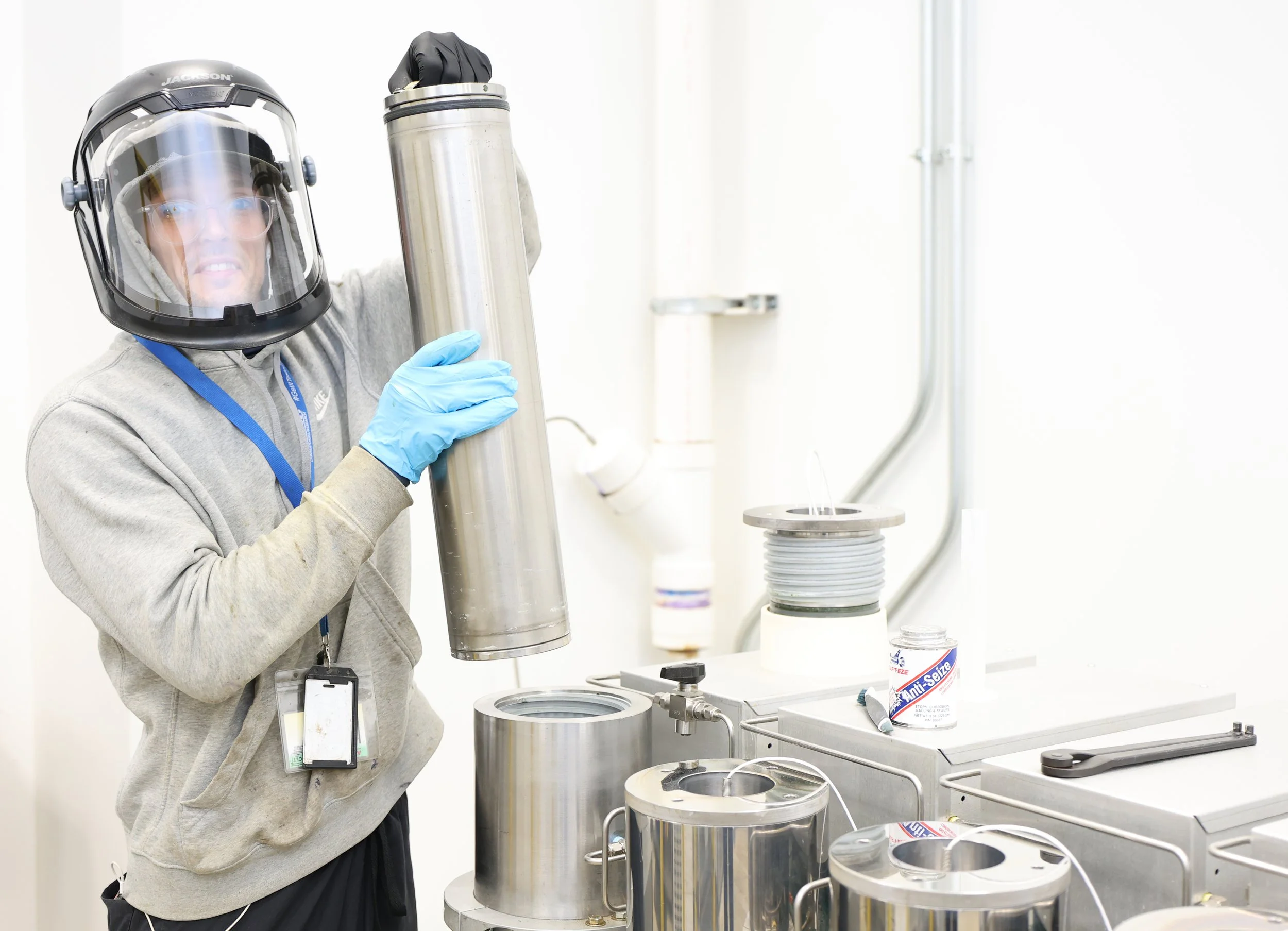 A scientist wearing protective gear, including a helmet with a face shield, gloves, and a hoodie, holding a large cylindrical metal container in a laboratory setting with various scientific equipment.