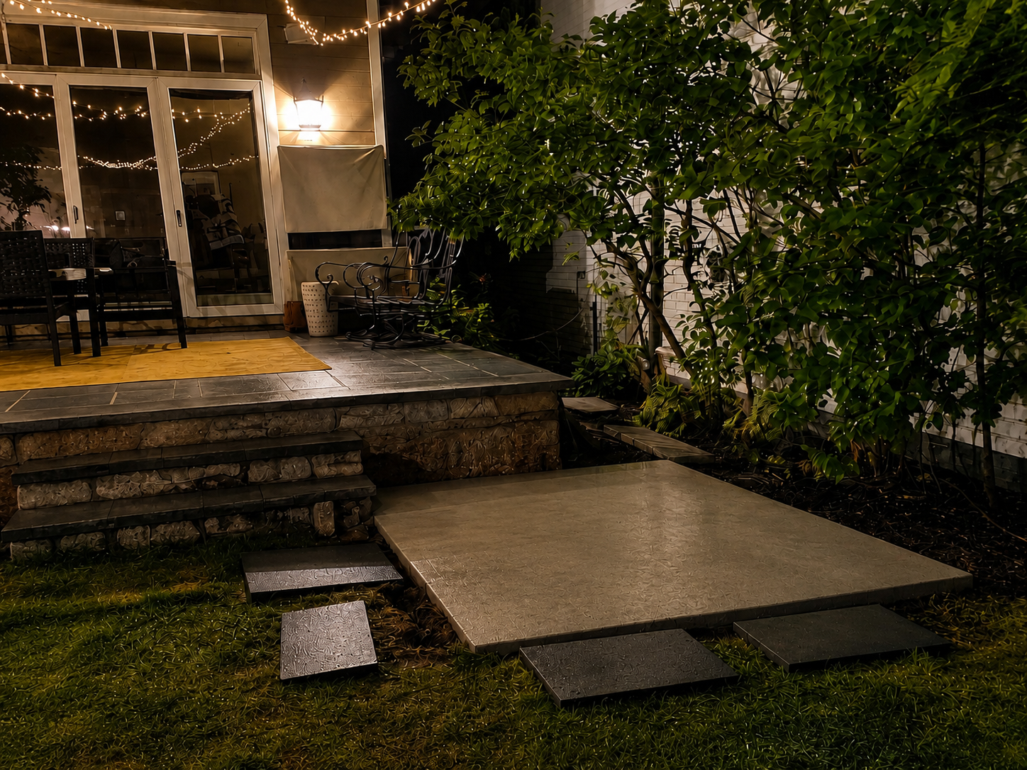 Nighttime view of a backyard patio with a small raised deck, a set of black chairs, a sliding glass door, string lights, green bushes, and a concrete stepping stone path.