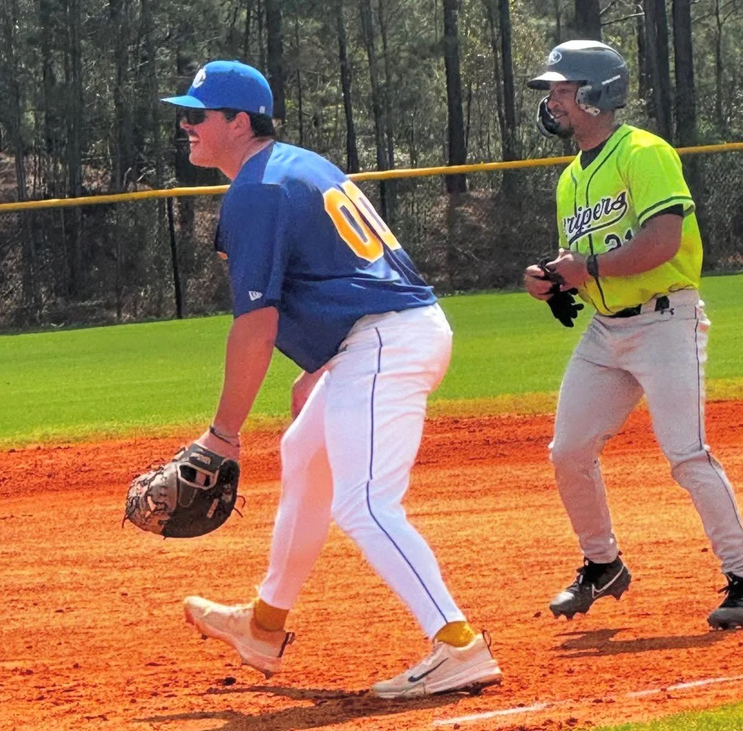 A baseball player in a blue uniform and cap is playing first base, while a baserunner in a yellow and black jersey and helmet is behind him, on a baseball field with green grass and trees in the background.
