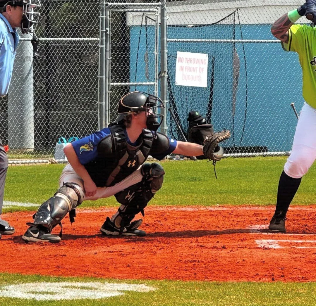 Baseball catcher in black protective gear crouching behind home plate, reaching out with his glove to catch a pitch, next to a batter in a bright yellow jersey, on a baseball field with a chain-link fence and a sign that says 'No Throwing in Front of