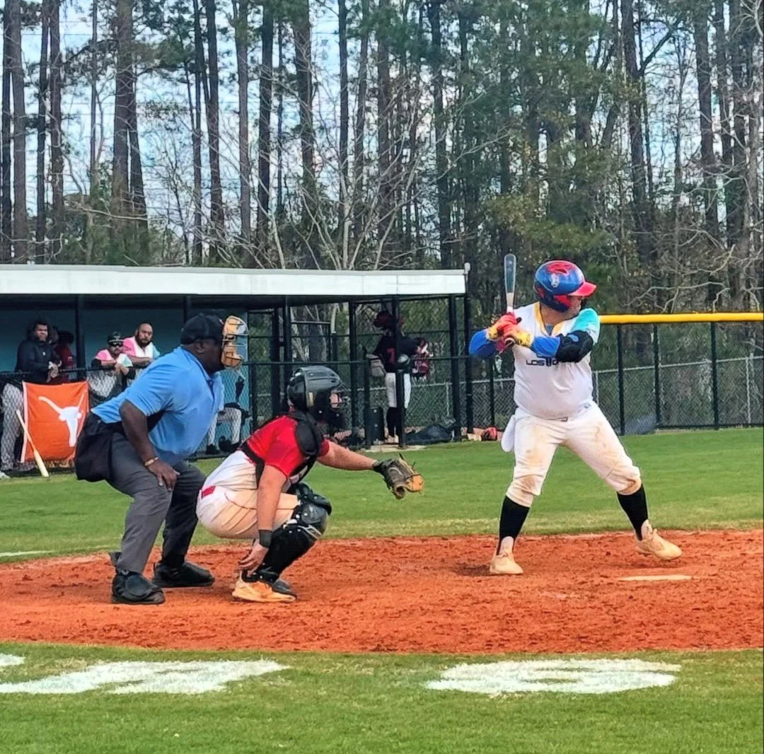 A baseball player at bat, catcher, and umpire during a game on a baseball field, with spectators and trees in the background.