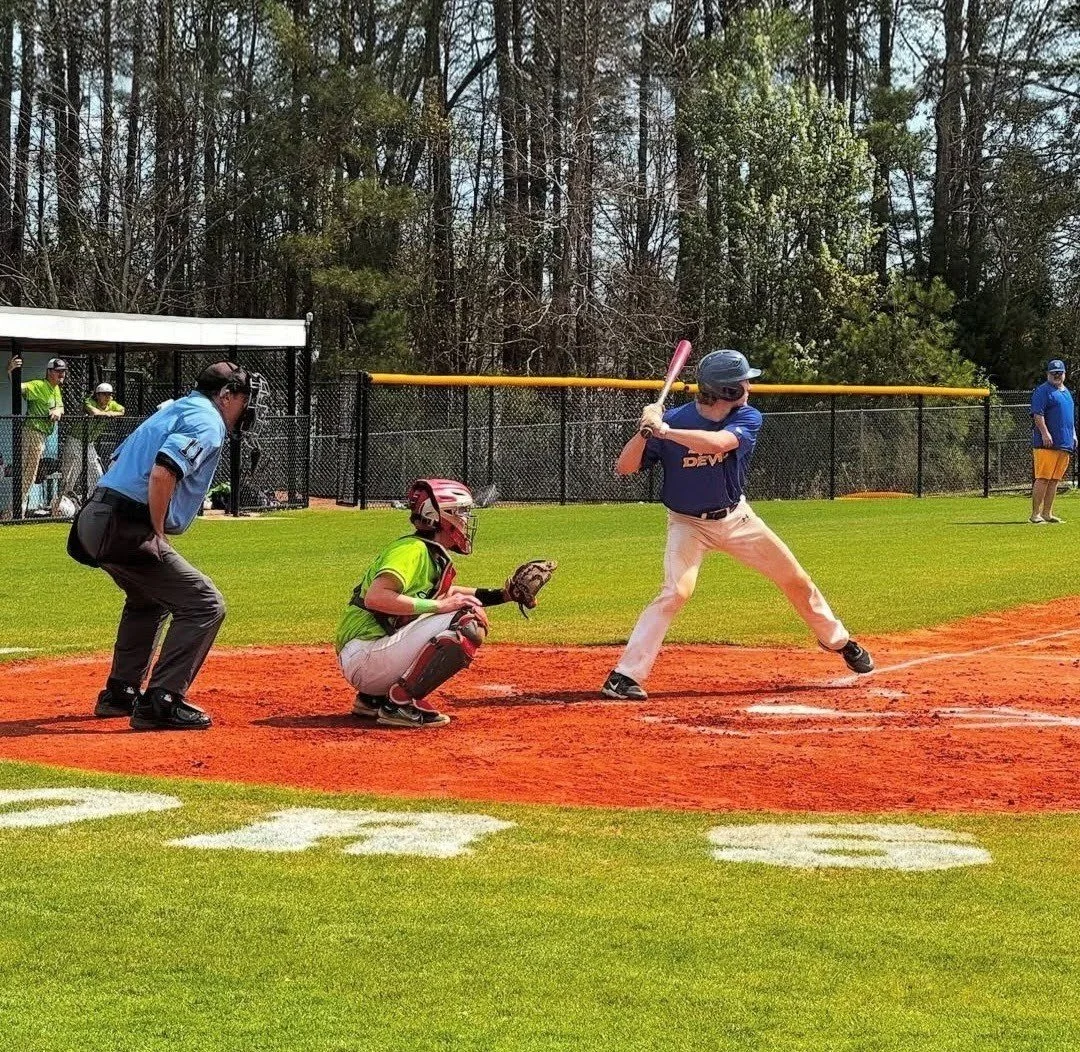 A baseball player swings at a pitch at home plate while the catcher and umpire prepare near the plate during a game on a sunny day. There are spectators behind a black fence and trees in the background.
