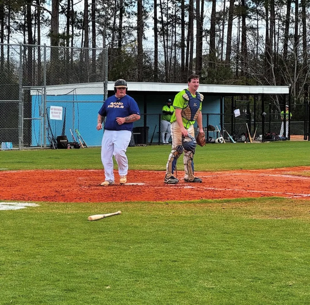 Two men standing on a baseball field, one in a blue shirt and white pants, the other in a green baseball catcher's gear, with a dugout and trees in the background.
