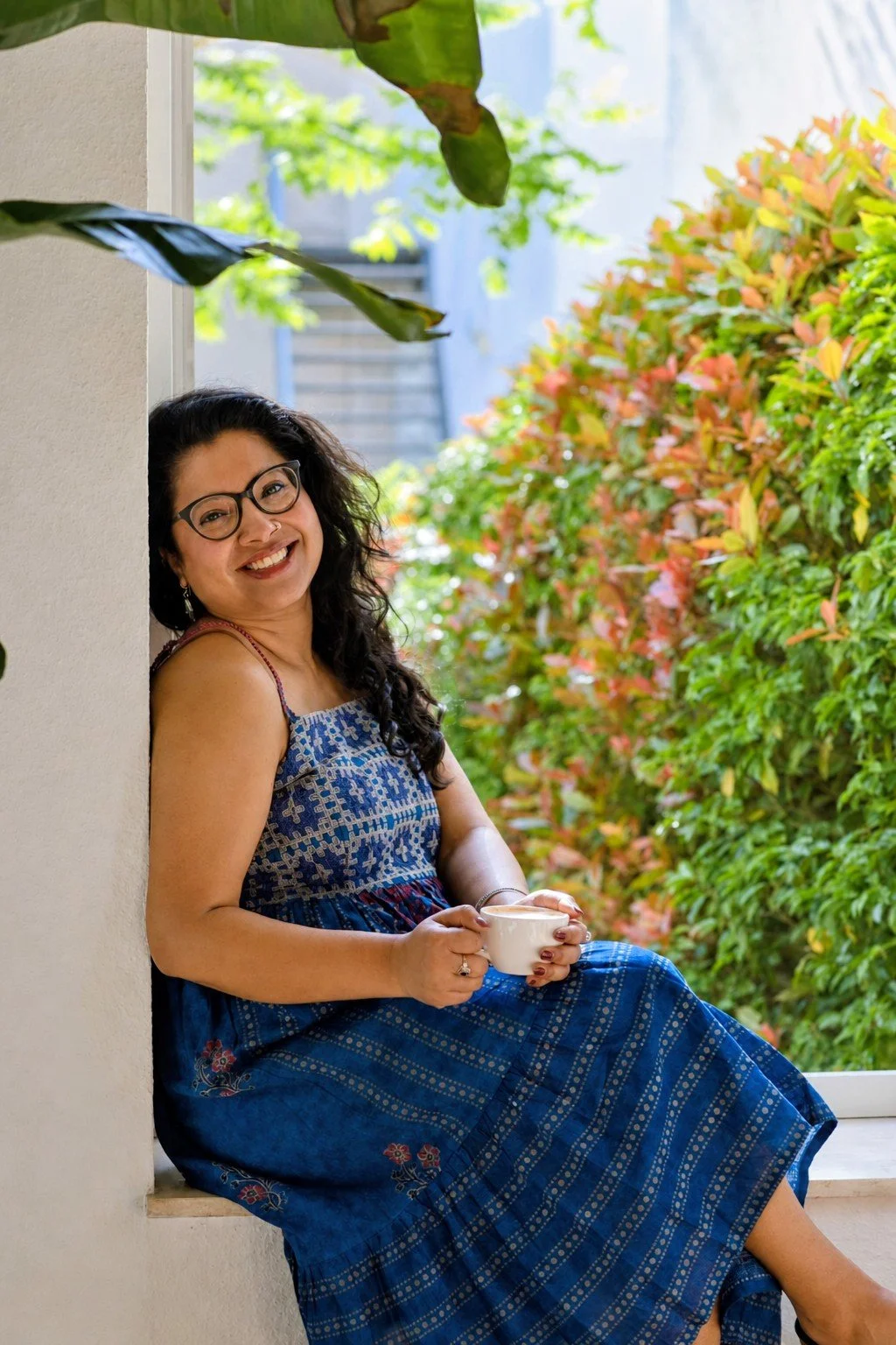 Dr. Pallabi Roy, sex, intimacy, & relationship coach, leaning against a window holding a coffee