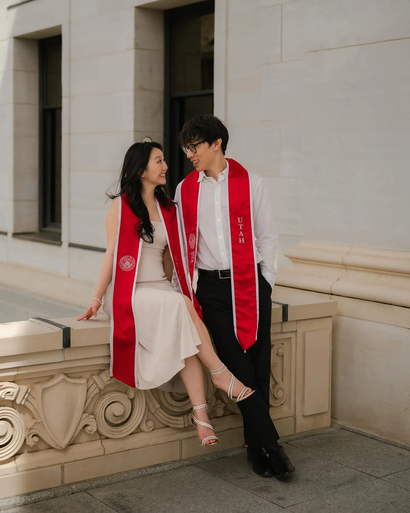 A man and woman dressed in graduation gowns, smiling and looking at each other. The woman is sitting on a decorative stone ledge, wearing a white dress and silver heels. The man is standing beside her, wearing a white shirt and black pants. Both are wearing red graduation sashes with the word 'Utah' printed on them.