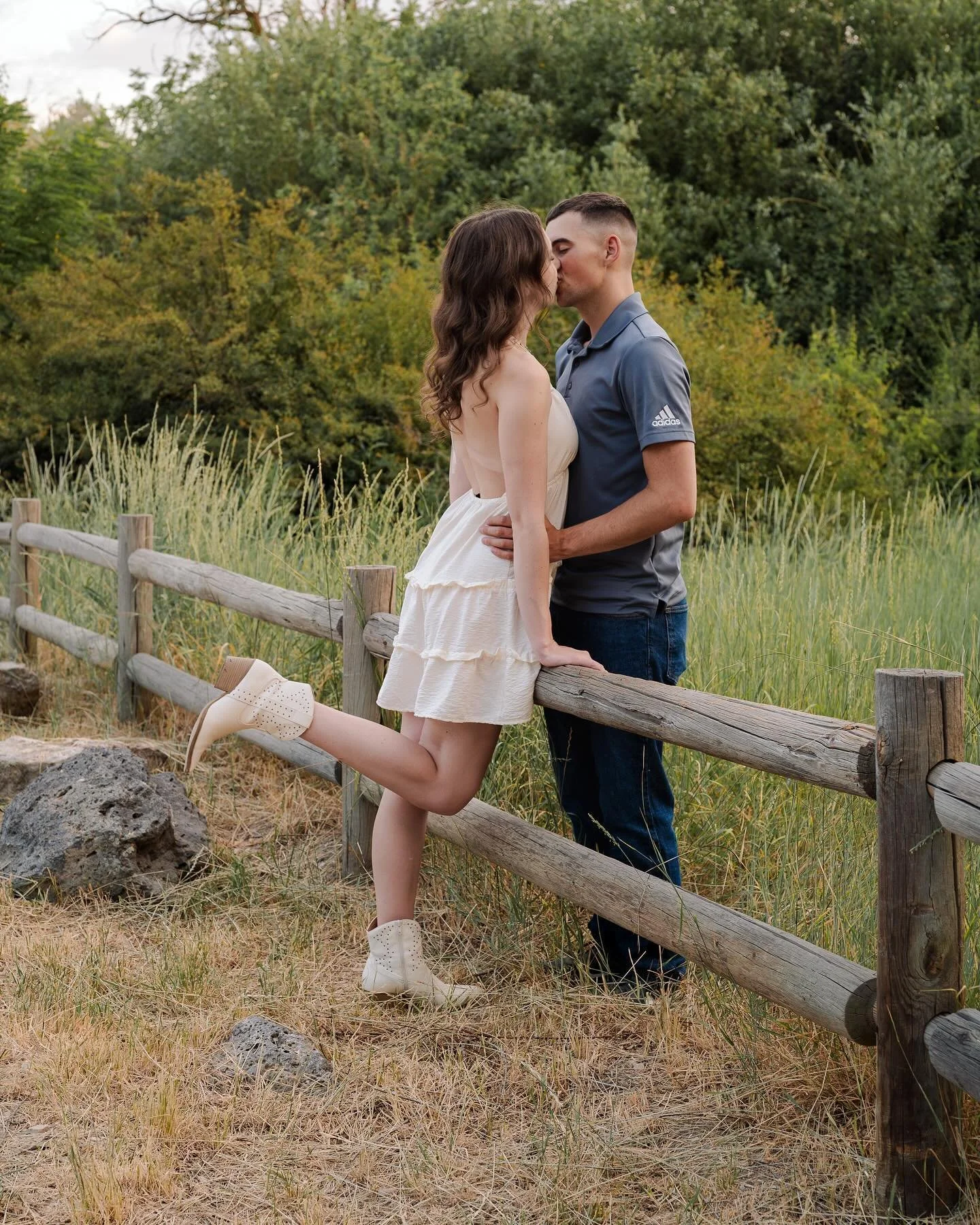 A couple kissing by a wooden fence in a grassy, wooded outdoor setting during the daytime.