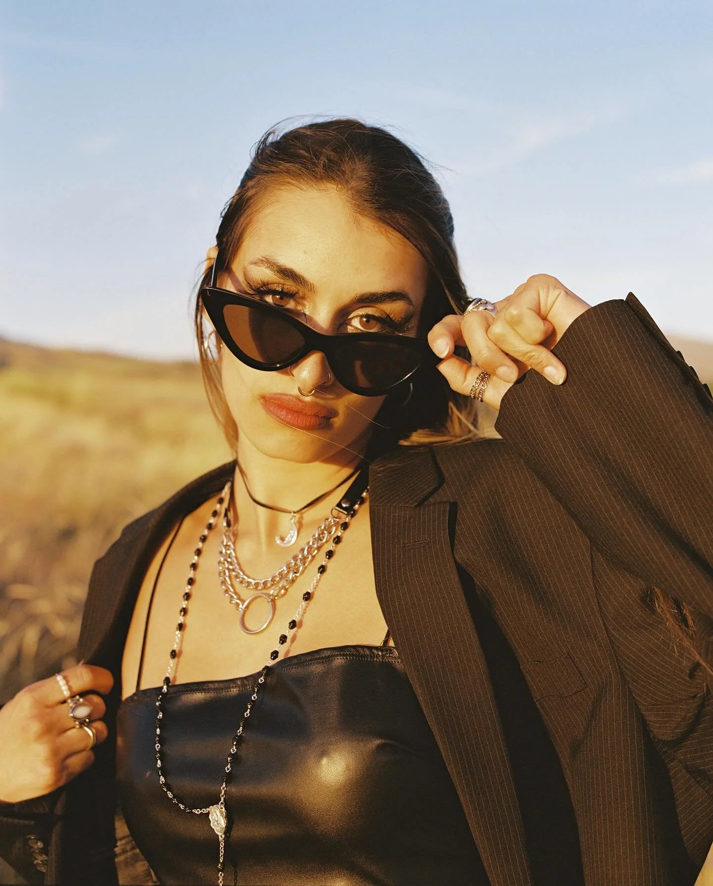 A woman with dark sunglasses, multiple necklaces, and rings on her fingers, holding the sunglasses with one hand, wearing a blazer over a black top, outdoors during sunset or golden hour.
