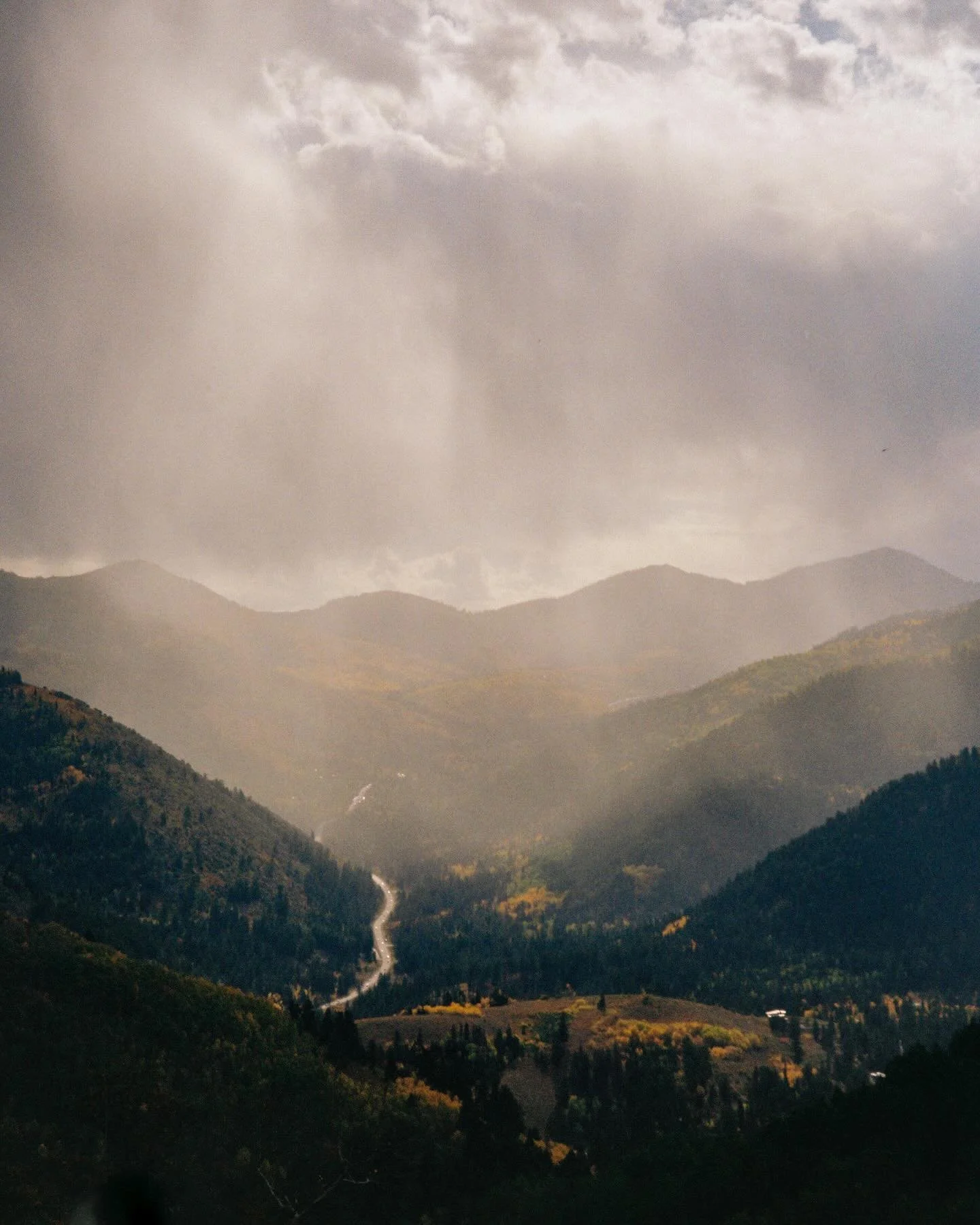 Mountain range with a cloudy sky, and a winding road through a forested valley.