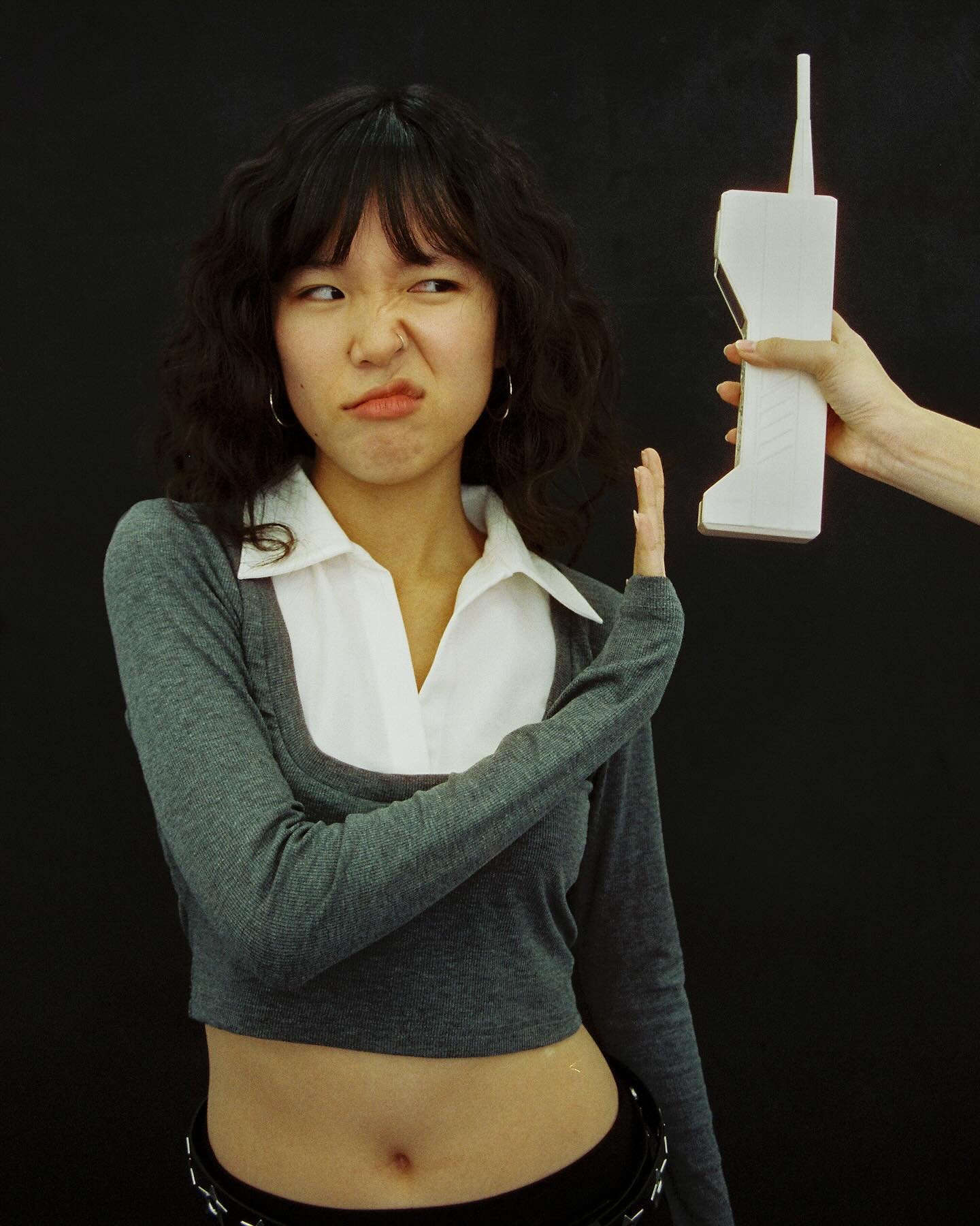 Young woman with curly dark hair making a disgusted face while holding up her hand away from a large white cordless phone being held toward her by a different person, against a black background.