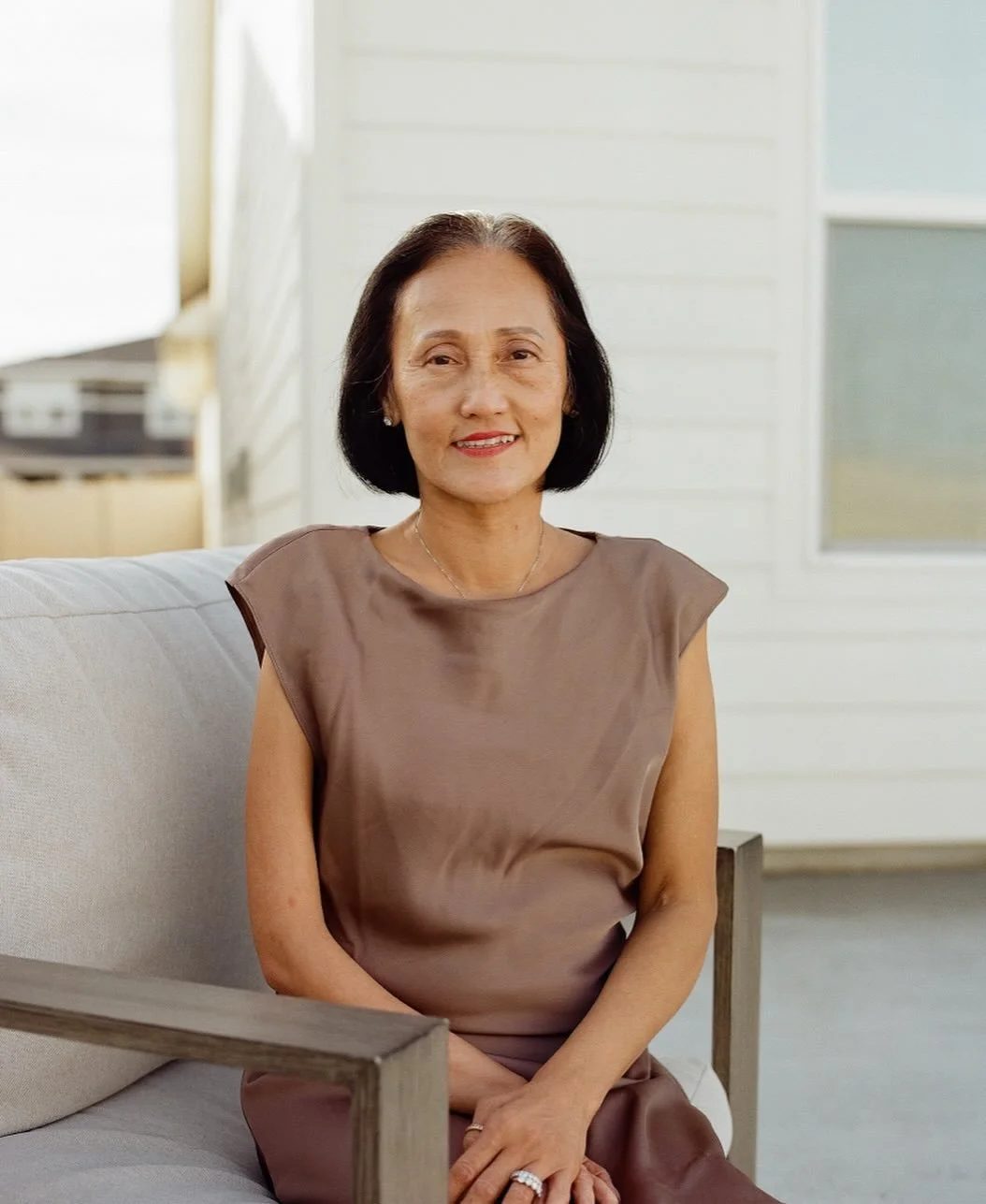 An elderly woman with short black hair sitting on a beige sofa in a bright room, smiling at the camera.