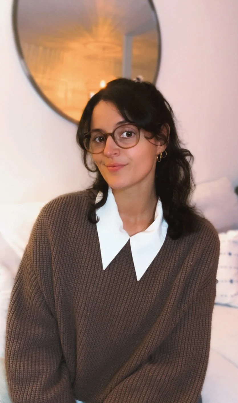 A young woman with dark, curly hair and glasses sitting indoors, wearing a brown sweater over a white collared shirt, in front of a round mirror reflecting warm lighting.