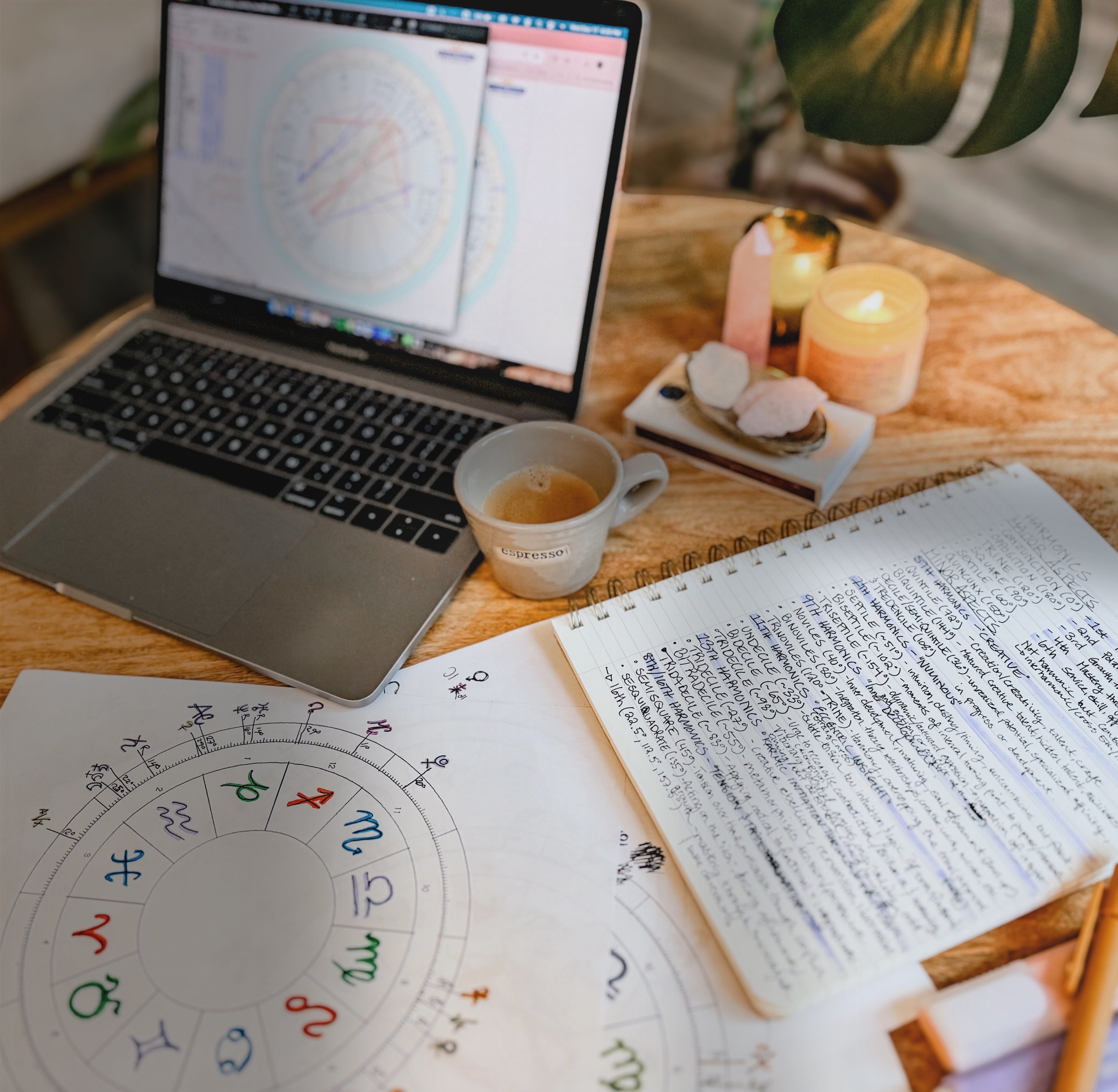 A workspace consisting of a MacBook laptop displaying a chart or diagram, an open notebook filled with handwritten notes, a printed circle chart with symbols representing zodiac signs, a cup of espresso, a candle, some crystals, and a pen on a wooden table.