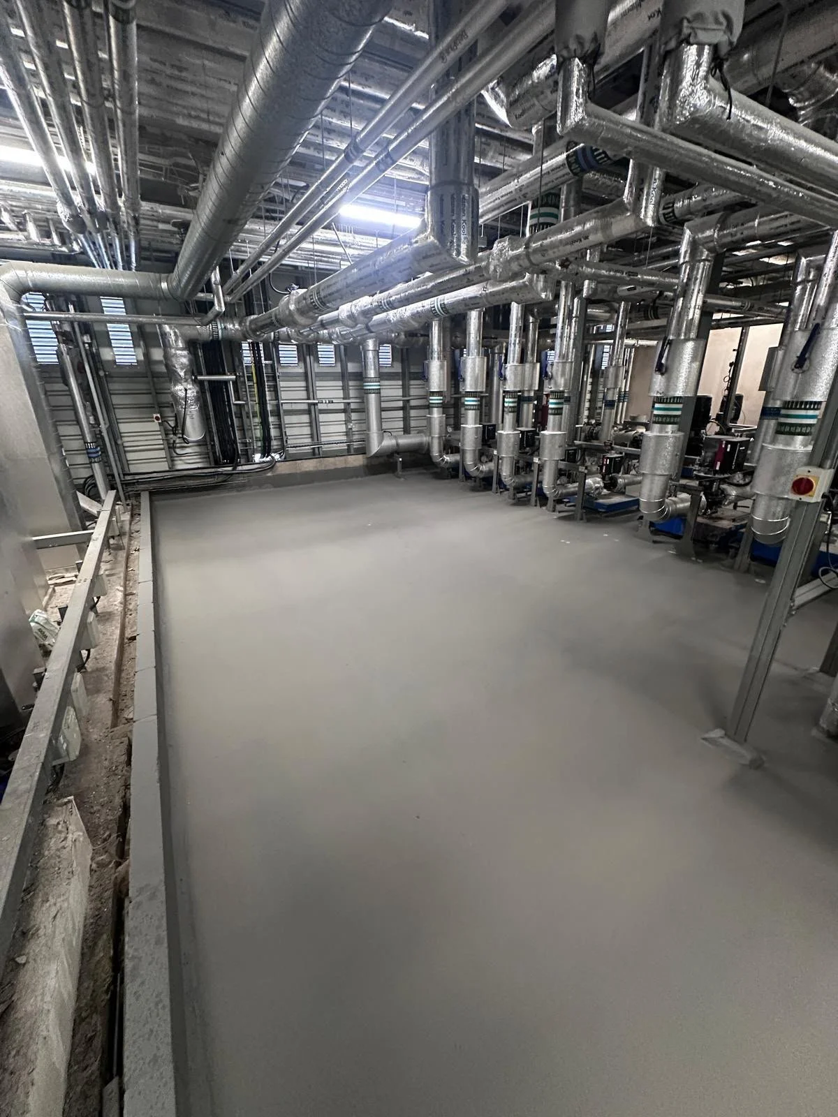 A room with metallic pipes and valves, likely part of an industrial or mechanical system, with a smooth concrete floor and some equipment along the right wall.
