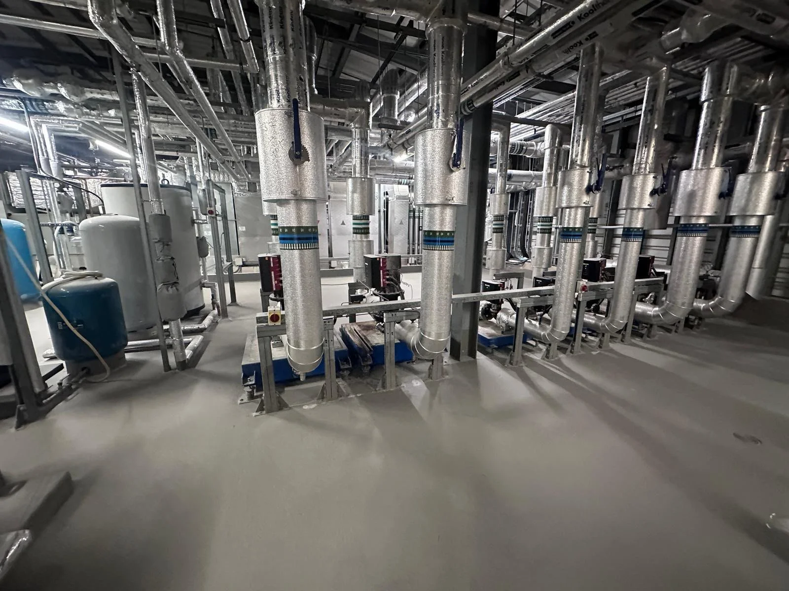 Industrial mechanical room with numerous silver pipes, blue and white tanks, and electrical components.