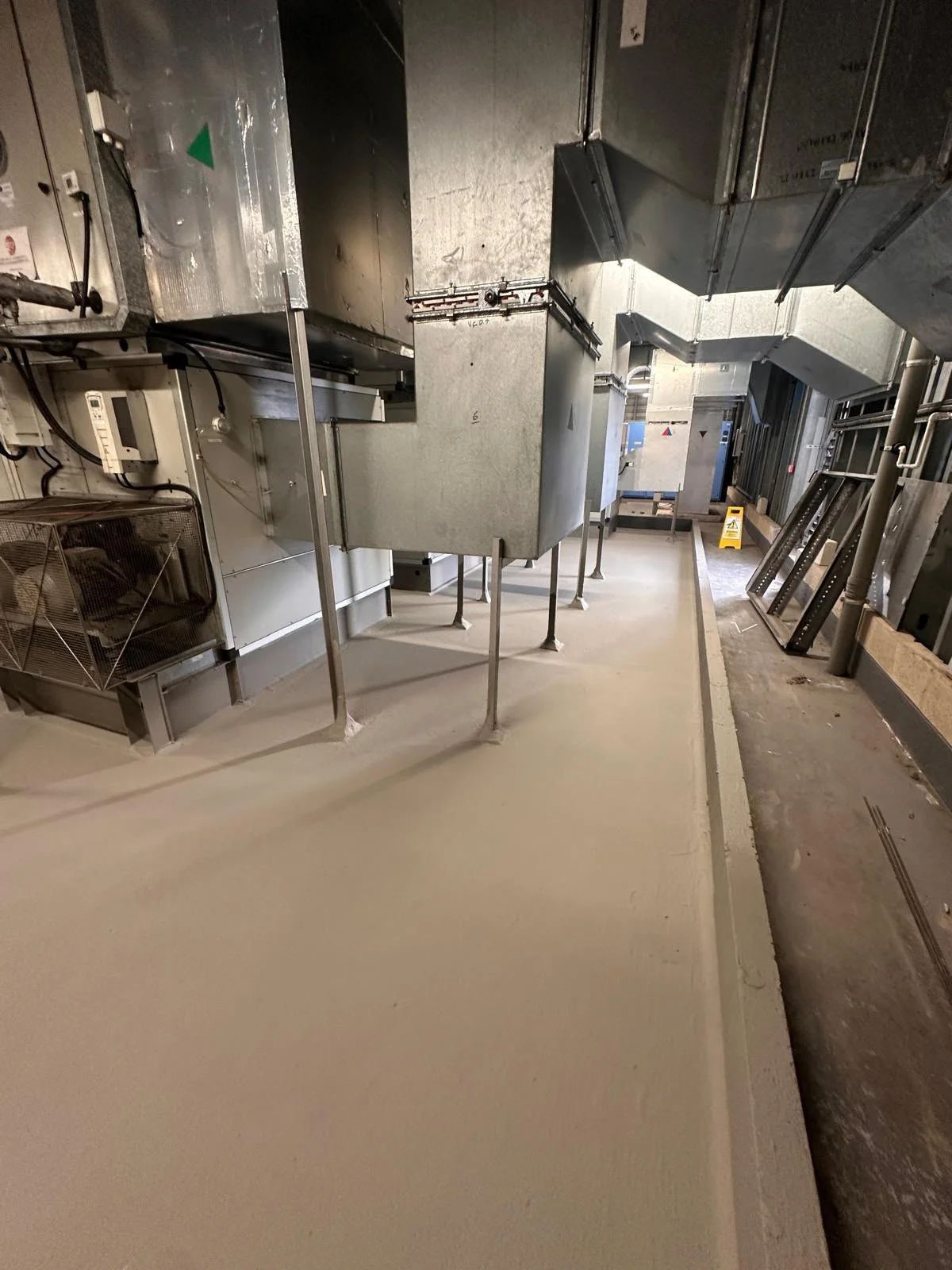 Interior view of a commercial or industrial kitchen or utility room with metallic ductwork, electrical panels, and a firm, clean floor, with some construction materials and a caution sign in the background.