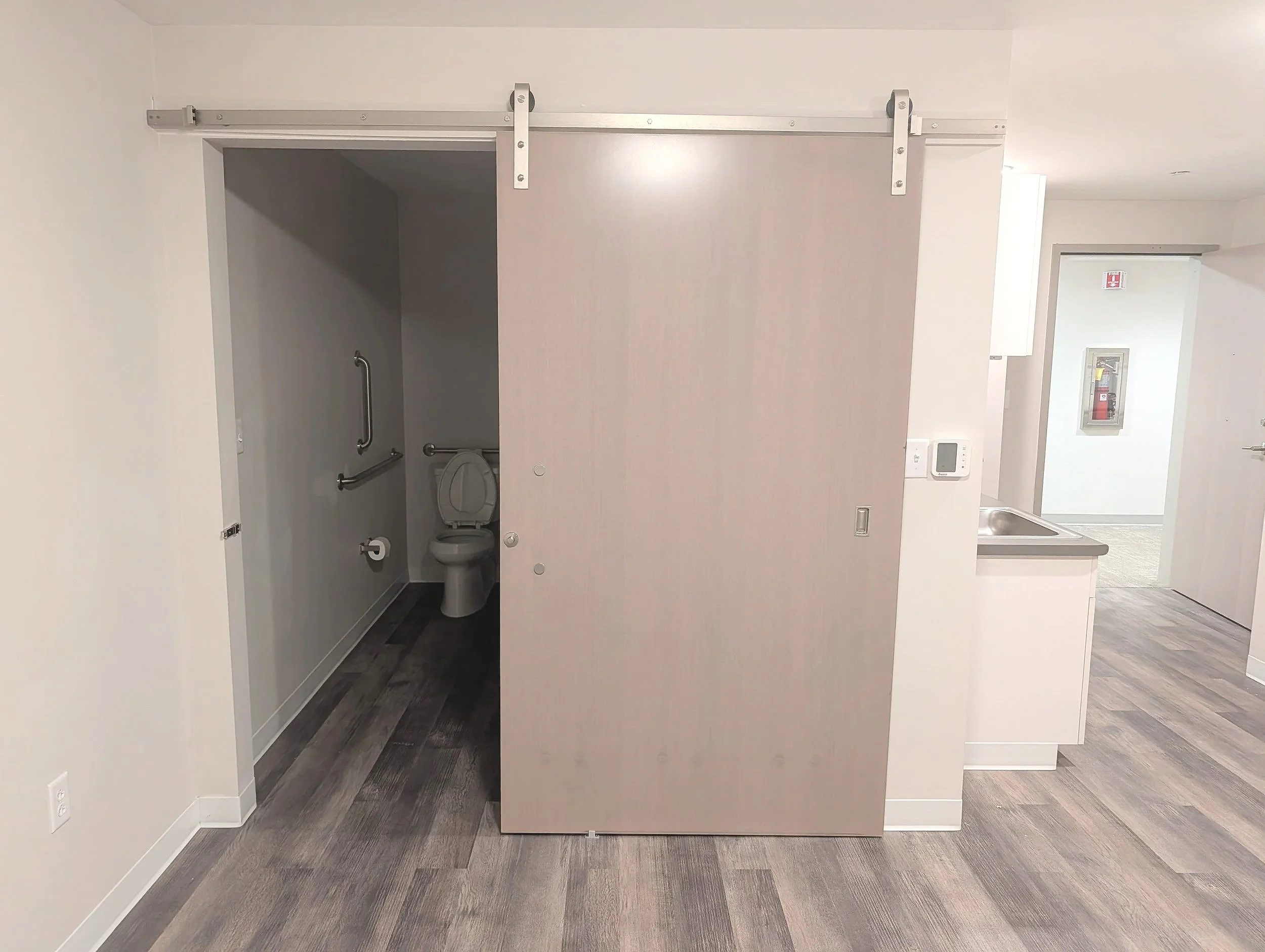 A sliding wooden door opens to reveal a small accessible restroom with a toilet and grab bars, situated next to a kitchenette area with a sink, in a modern building interior.