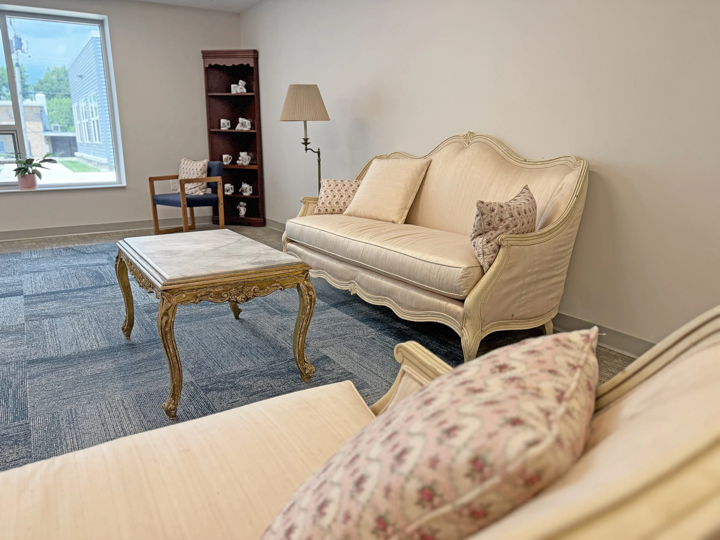 Living room with vintage cream-colored sofa, floral and beige throw pillows, a carved wooden coffee table, a corner bookcase with china, a blue armchair, a window with a potted plant, and a tall floor lamp.
