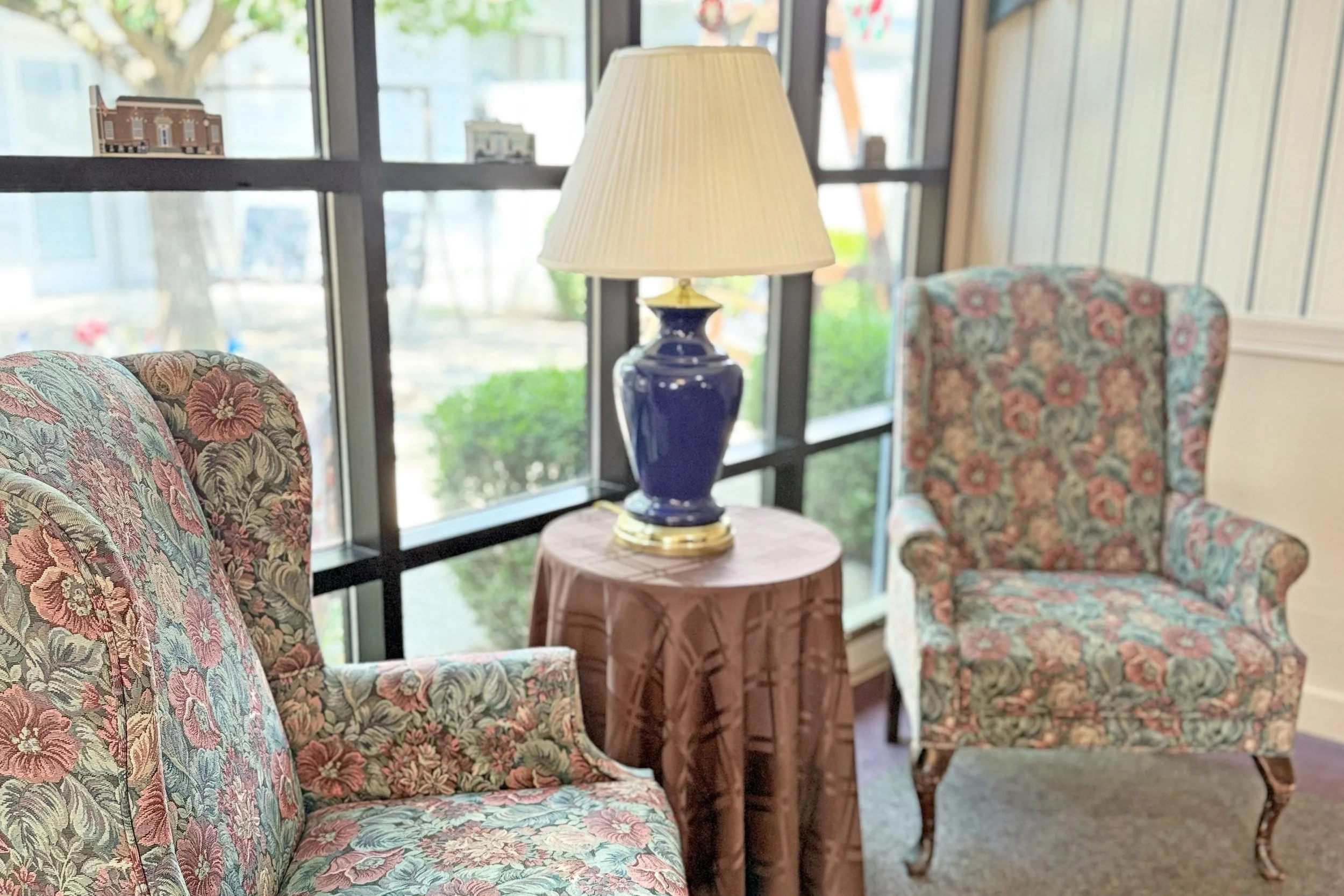 Sitting area with two floral armchairs, a side table with a large blue and gold lamp, and large windows in the background with trees and buildings visible outside.
