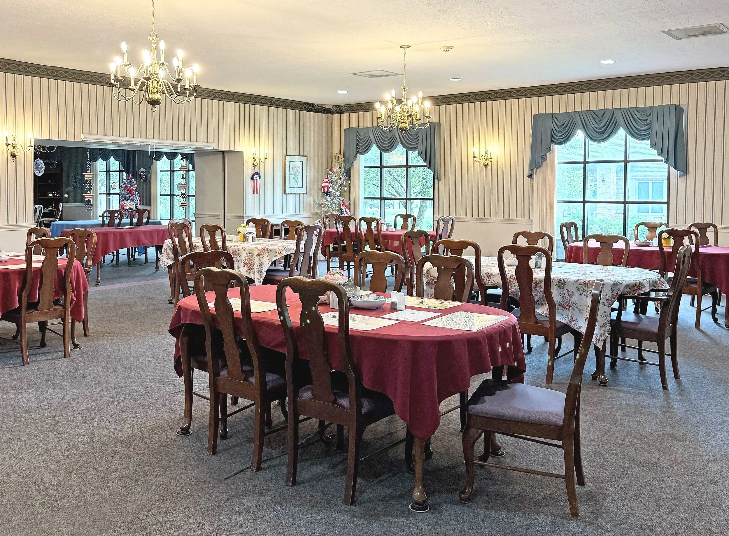 Dining room decorated for Christmas with tables covered in red and floral tablecloths, wooden chairs, large windows with blue curtains, and a Christmas tree with ornaments in the background.
