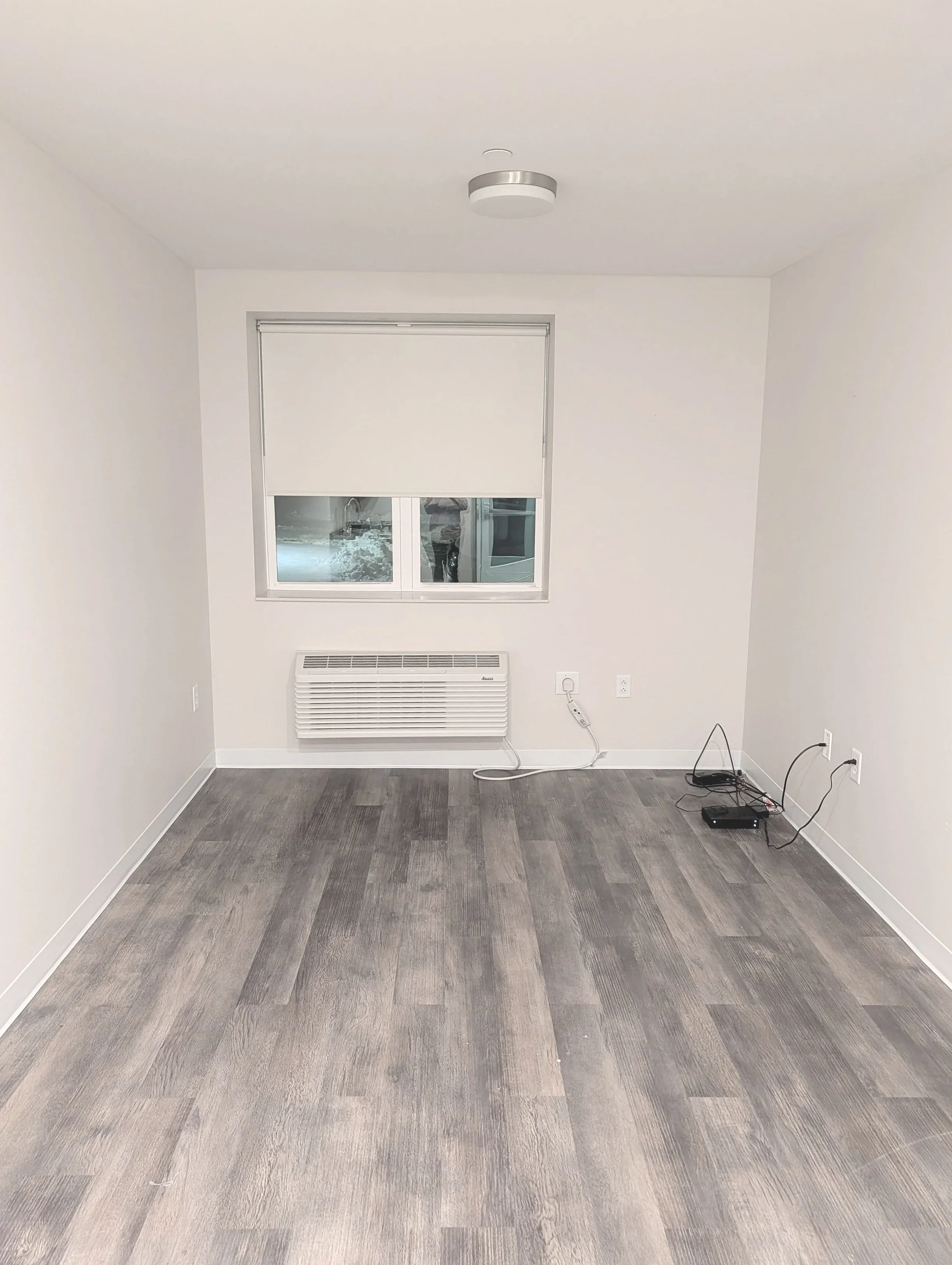 Empty room with gray wood flooring, white walls, a window with a closed white blind, a wall-mounted heater, and some electronic devices and wires on the floor.
