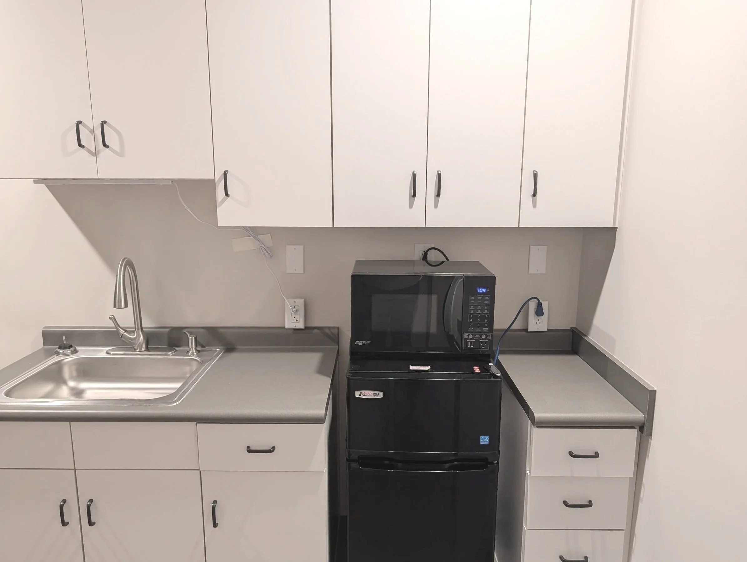 A compact kitchen with white cabinets, a stainless steel sink, a microwave oven on top of a black mini refrigerator, and a countertop with multiple drawers.