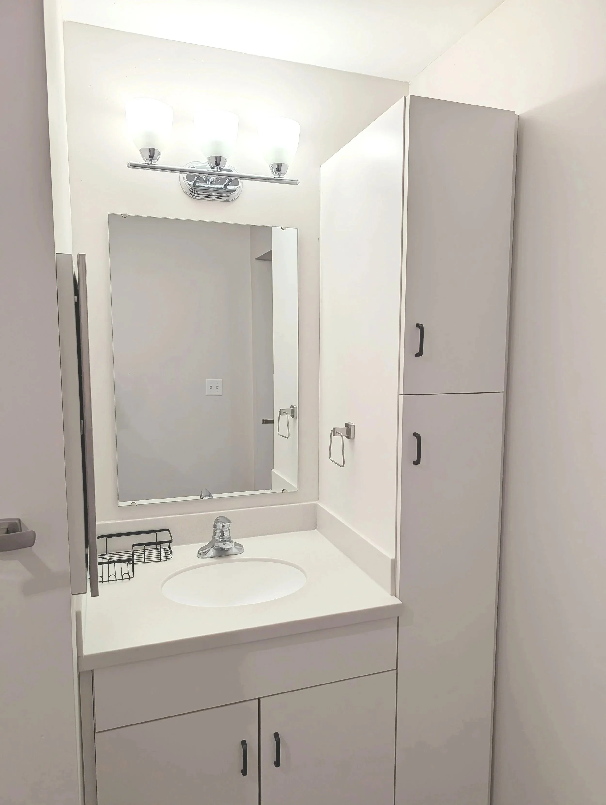 A bathroom vanity with a white countertop, oval sink, single-handle faucet, mirror, wall-mounted light fixture, and storage cabinet with silver handles.