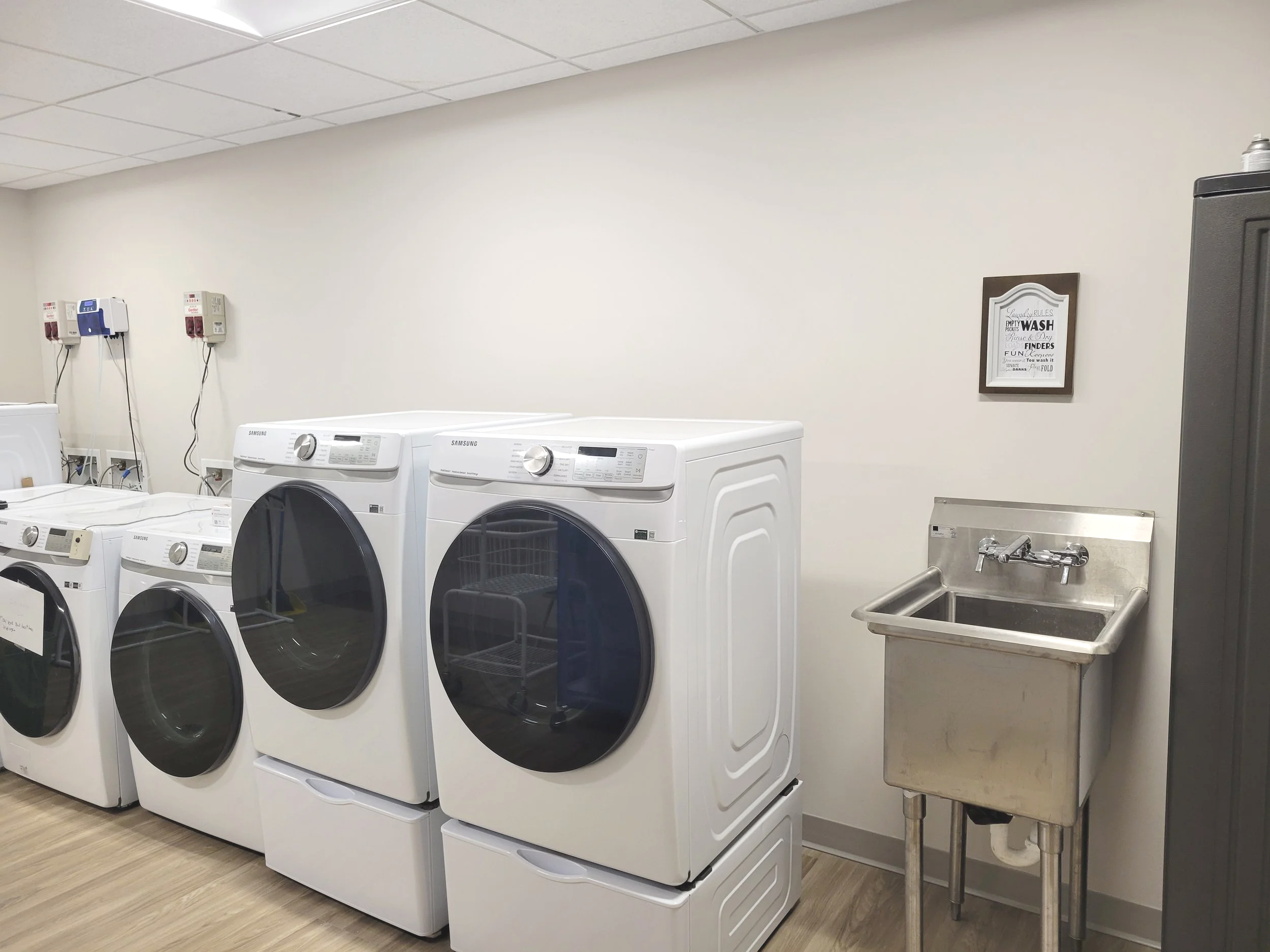 A laundry room with three front-loading washing machines, a stainless steel utility sink, and a framed sign on the wall.