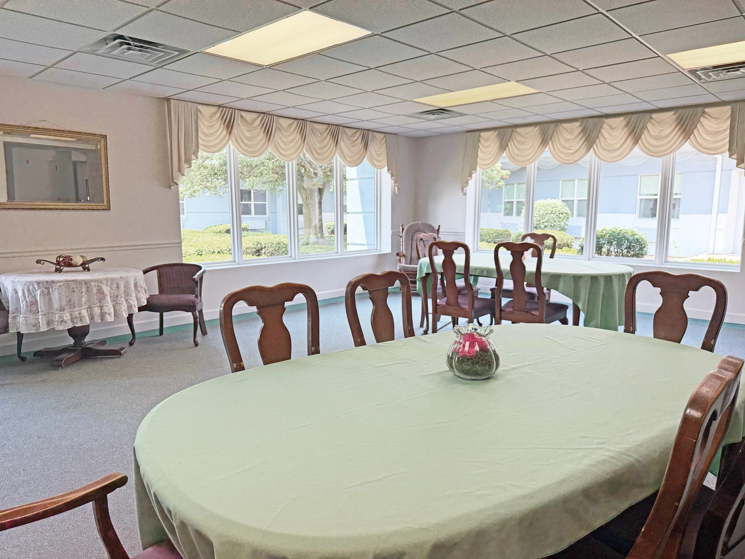 A dining room with multiple tables covered with light green and green tablecloths, surrounded by wooden chairs, and decorated with a small glass vase with pink flowers. The room has large windows with cream-colored drapes, letting in natural light, with a view of trees and a building outside.