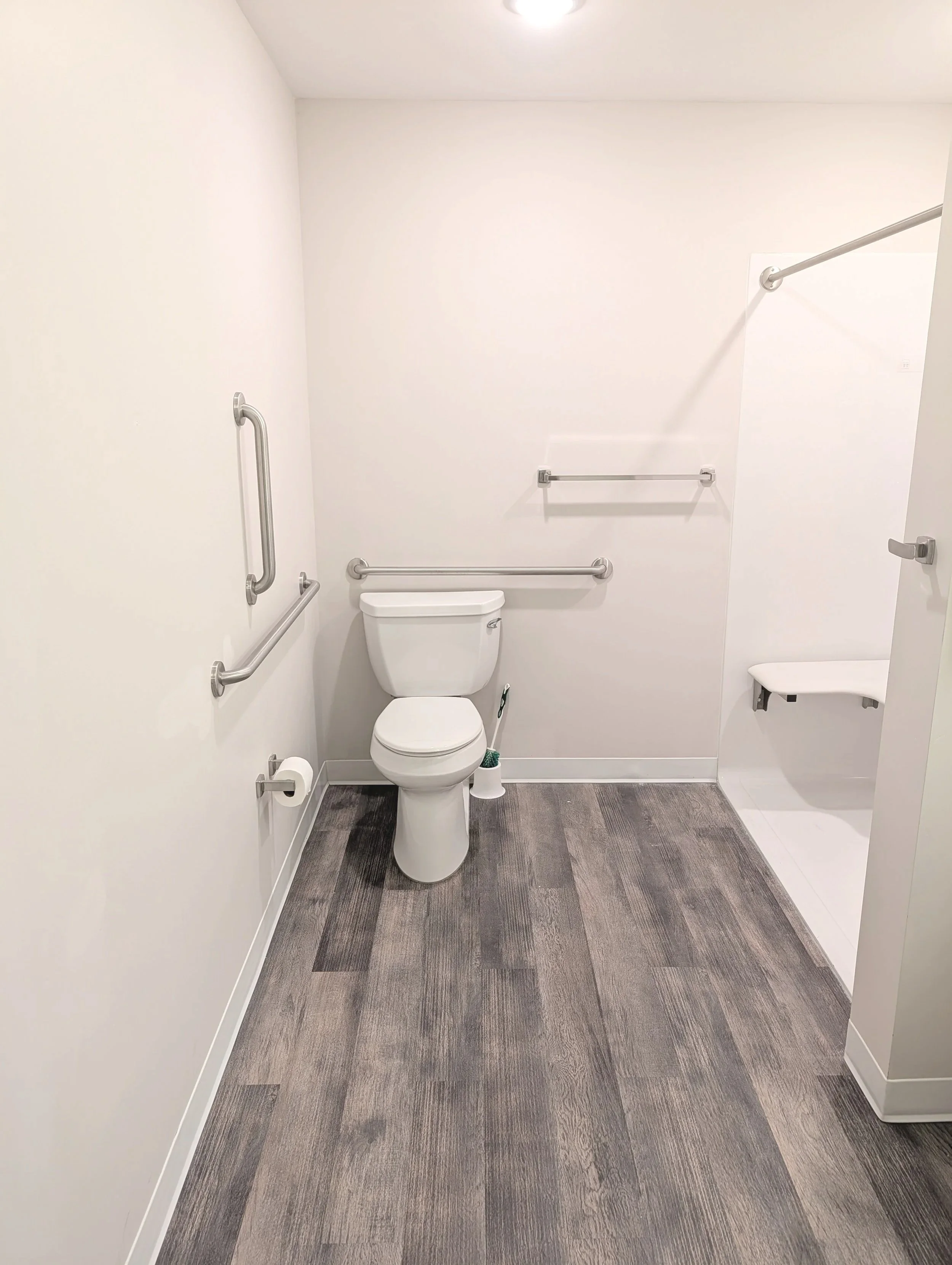 Accessible bathroom with white walls, gray wood-style flooring, a toilet, grab bars, a white shelf, and a small bench in the shower area.