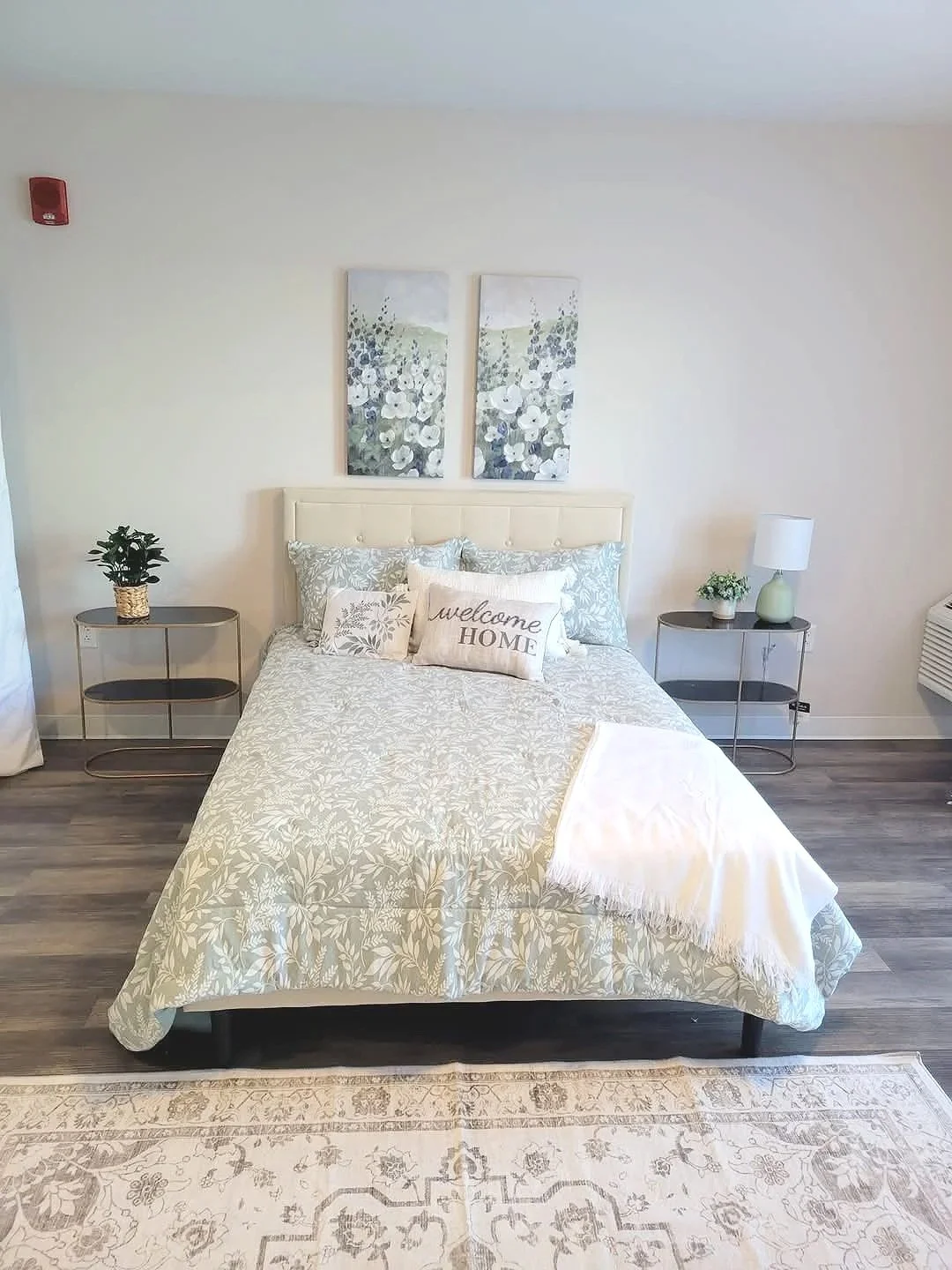 A bedroom with a bed, two nightstands, and decorative wall art. The bed has a headboard, patterned bedding, and multiple pillows including one with 'welcome HOME' embroidered. There are potted plants on each nightstand, a lamp on the right, and a framed artwork of floral scenery above the bed. The floor is wood, and there's a patterned rug at the foot of the bed.
