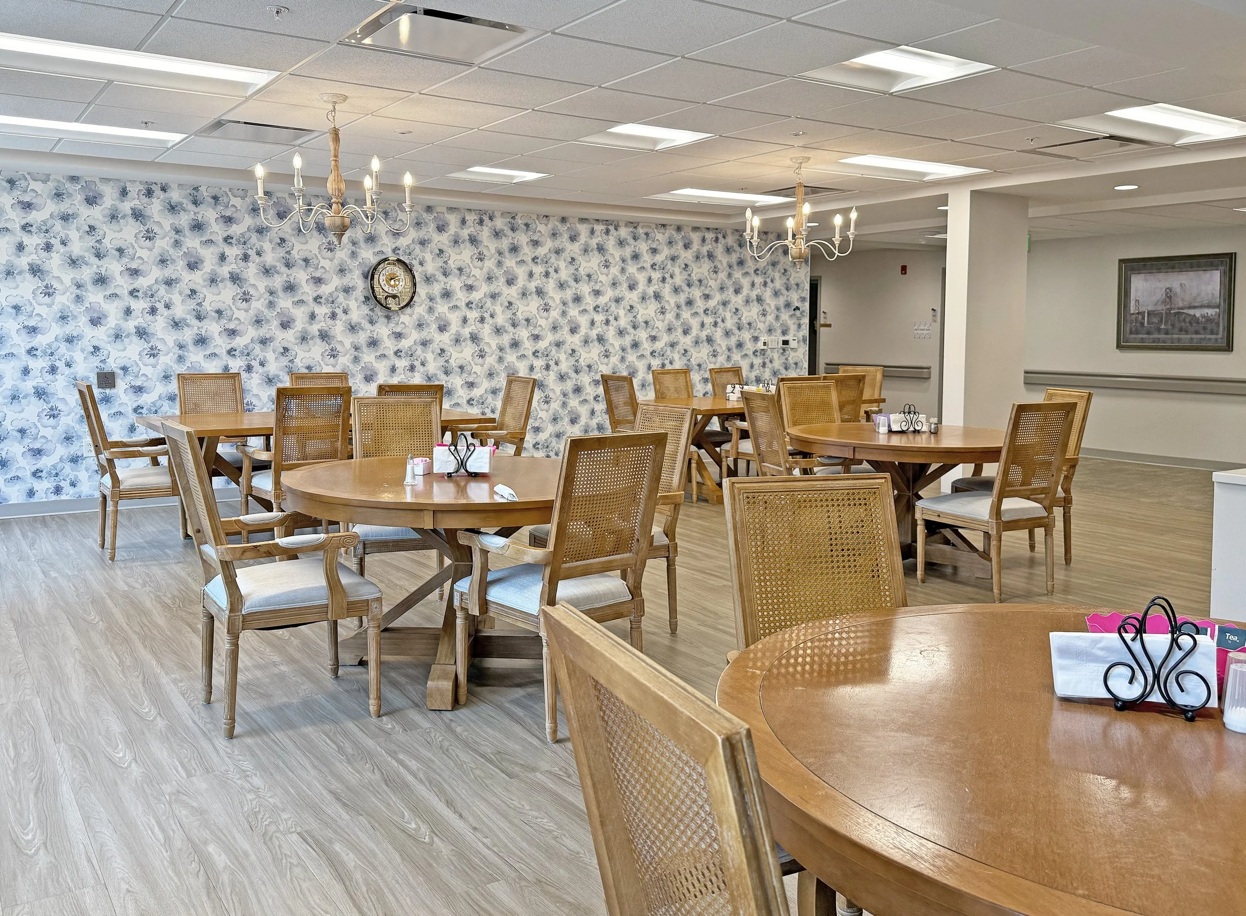 Empty dining area with wooden tables and chairs, floral wallpaper on one wall, chandeliers hanging from the ceiling, and a framed picture on the wall.