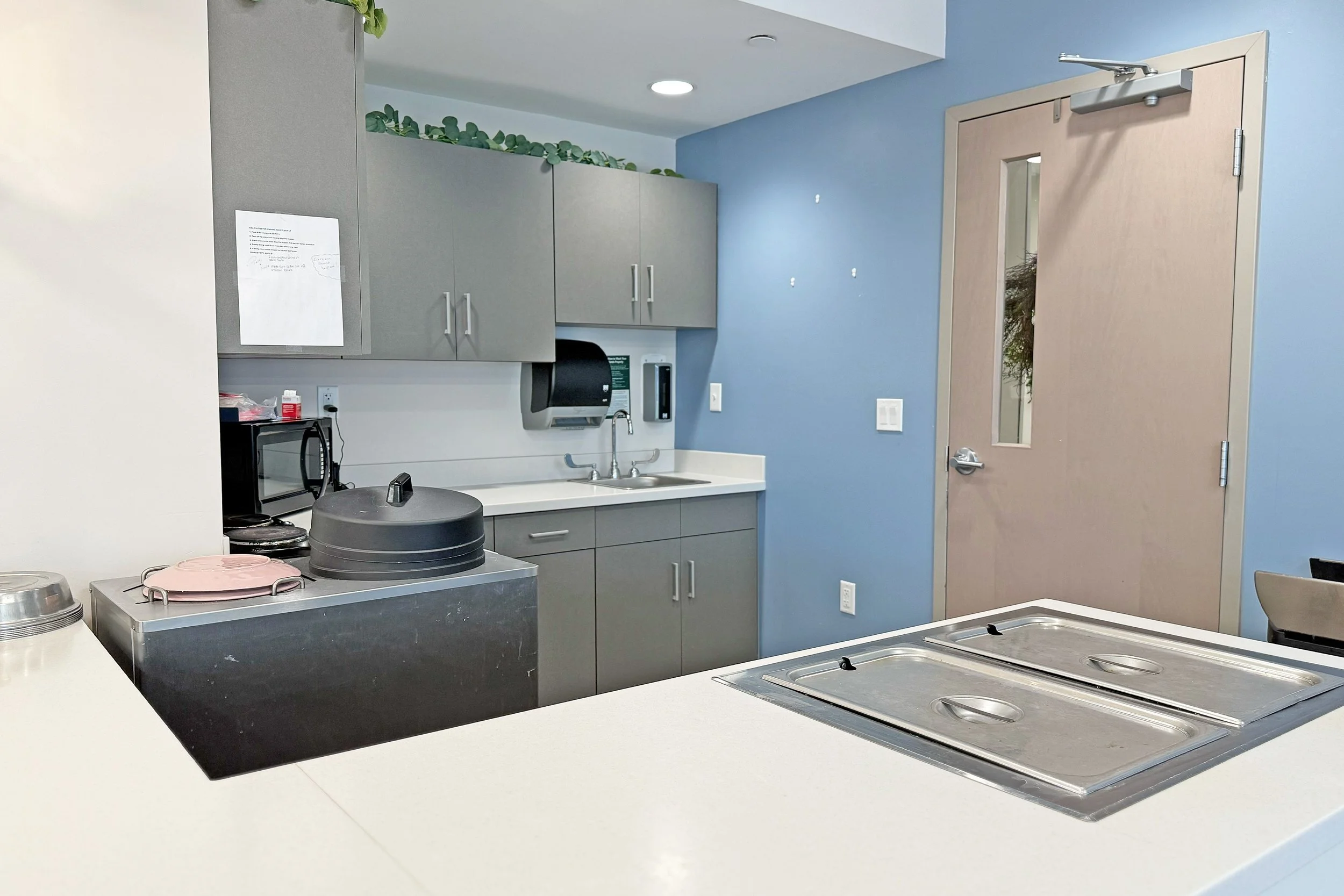 Hospital or medical clinic kitchen area with gray cabinets, a blue wall, a door with a small window, and a counter with a metal food service cart and covered trays.