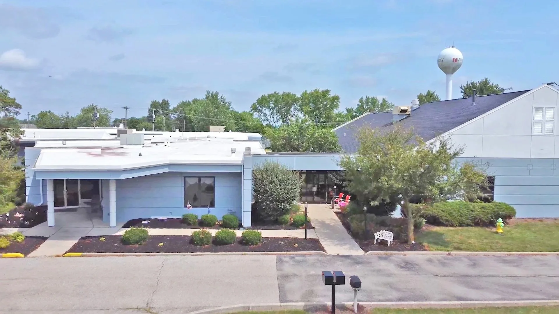A daytime view of a light blue building with a front porch, surrounded by landscaped bushes and trees. There is a sidewalk leading to the entrance with white chairs outside, and a parking area in front. In the background, there is a water tower and some greenery.