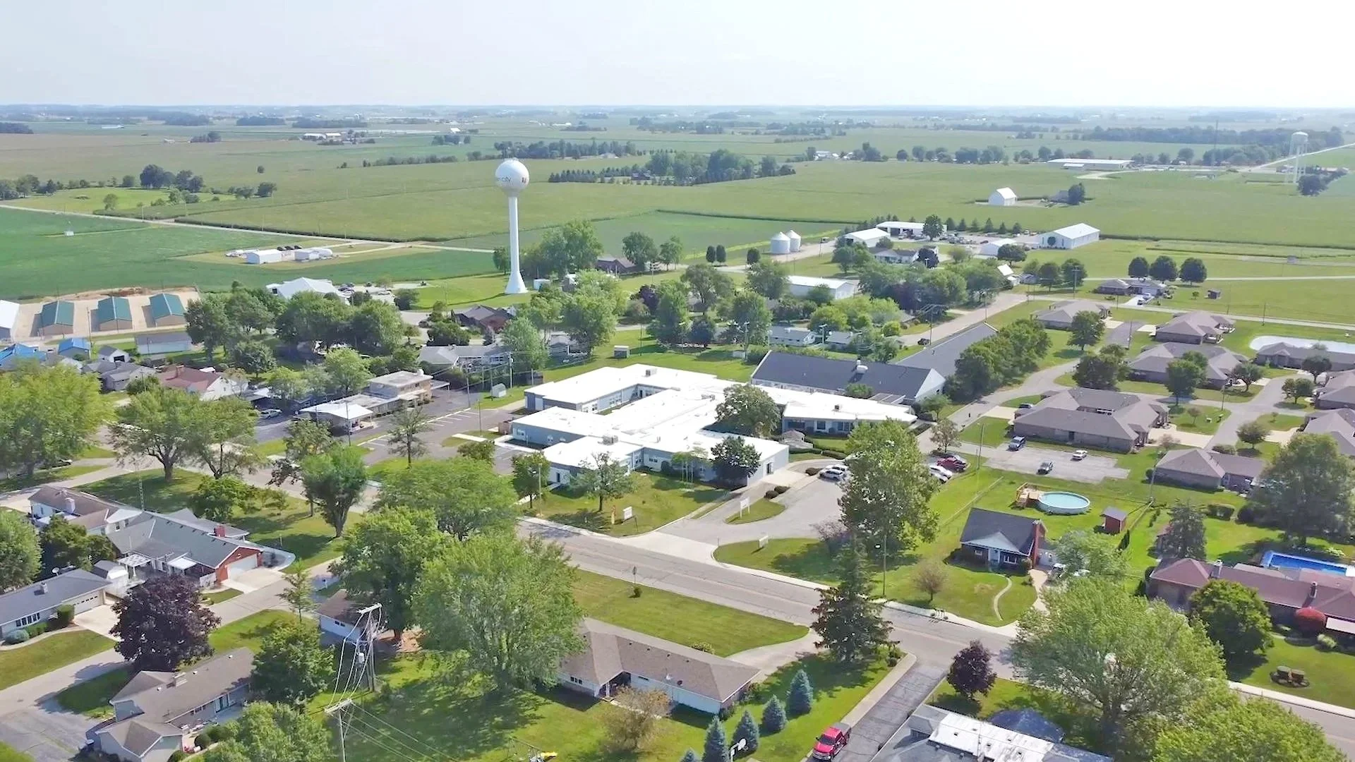 Aerial view of a small town surrounded by green fields and farmland, with houses, trees, and a water tower.