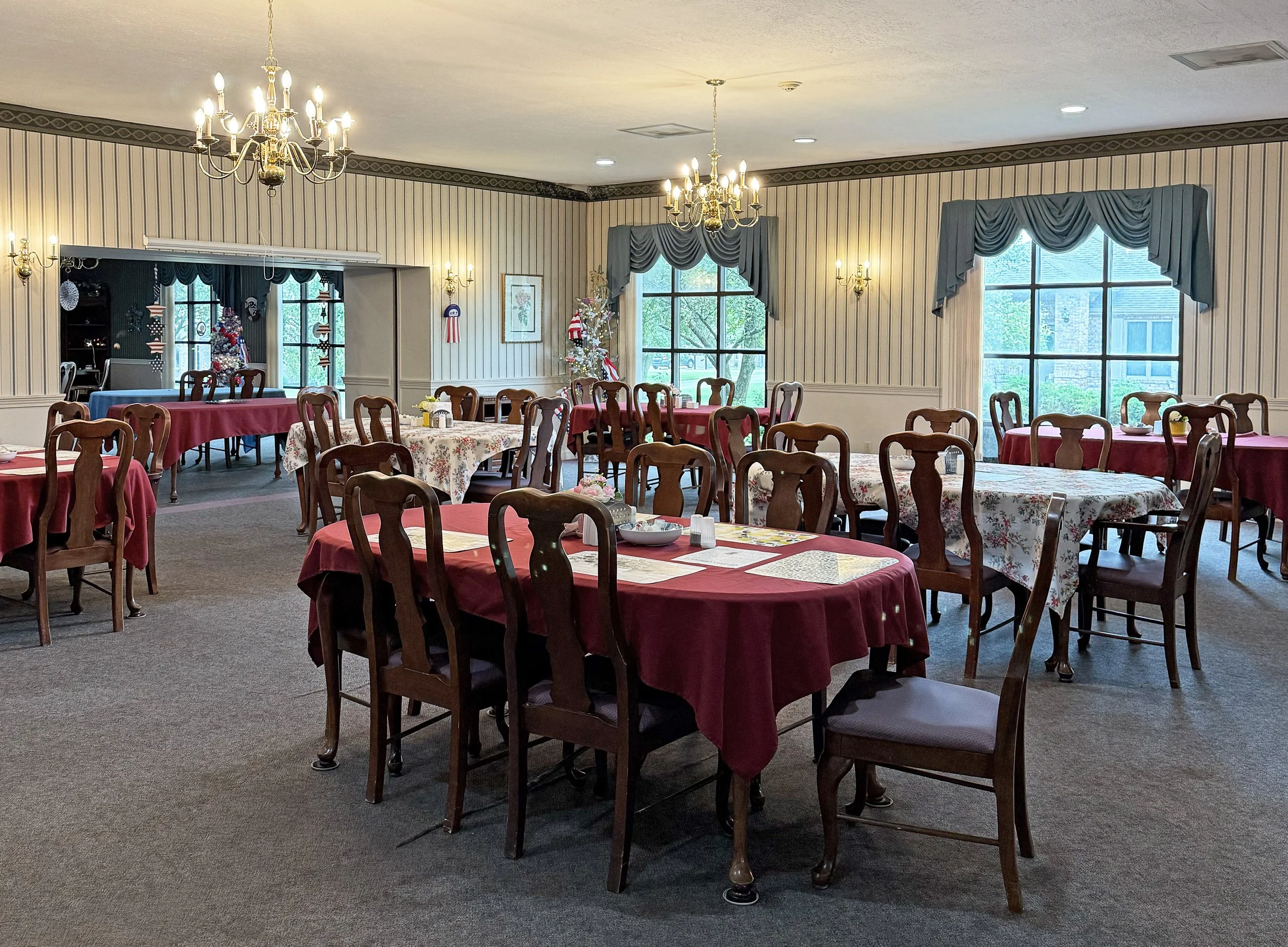 Elegant dining room decorated for Christmas with wooden tables, red and floral tablecloths, wooden chairs, chandeliers, and windows with blue curtains.